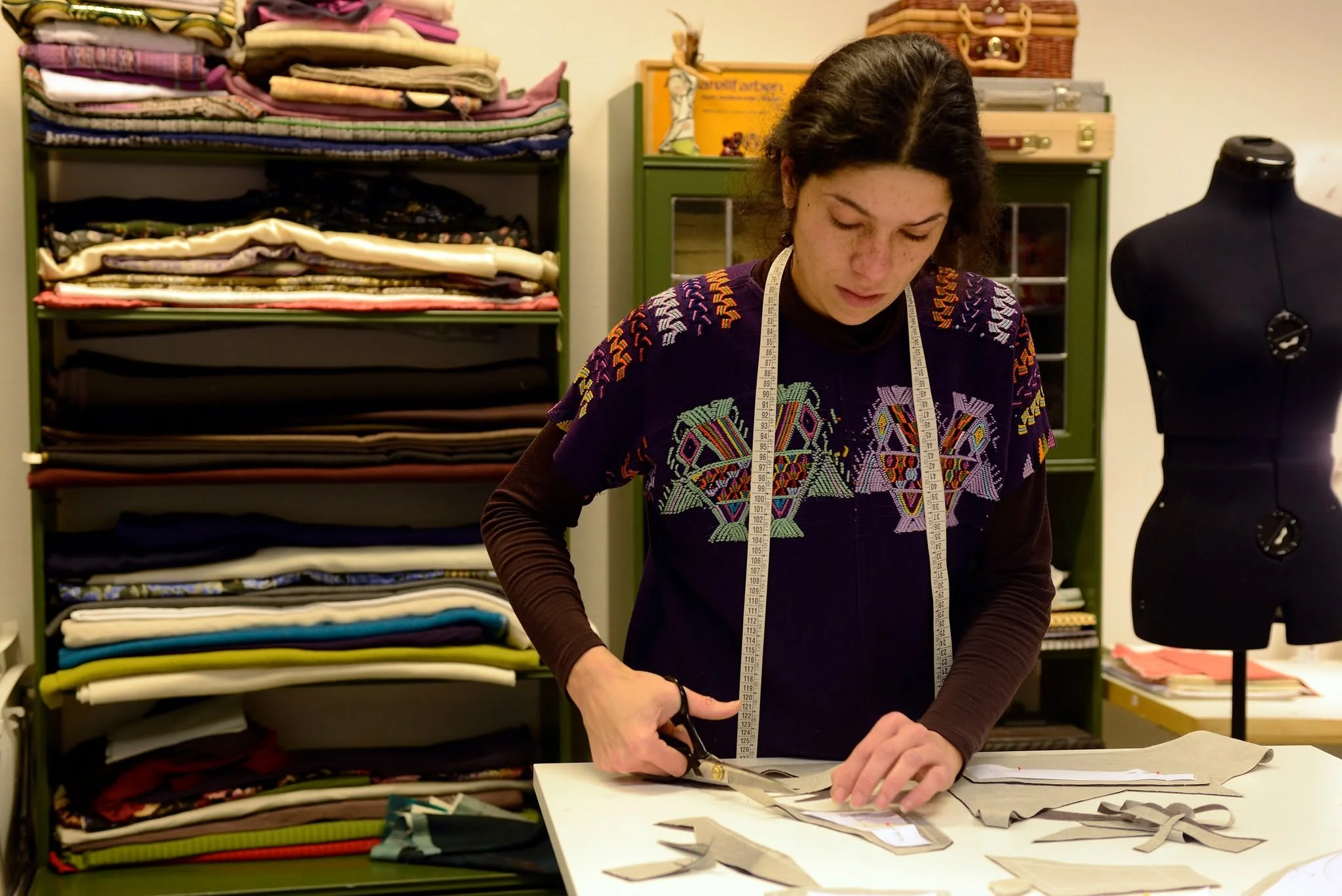 A woman working with fabric and scissors at a table in a fabric store or workshop, with shelves of colorful fabric behind her and a dress form on her right.