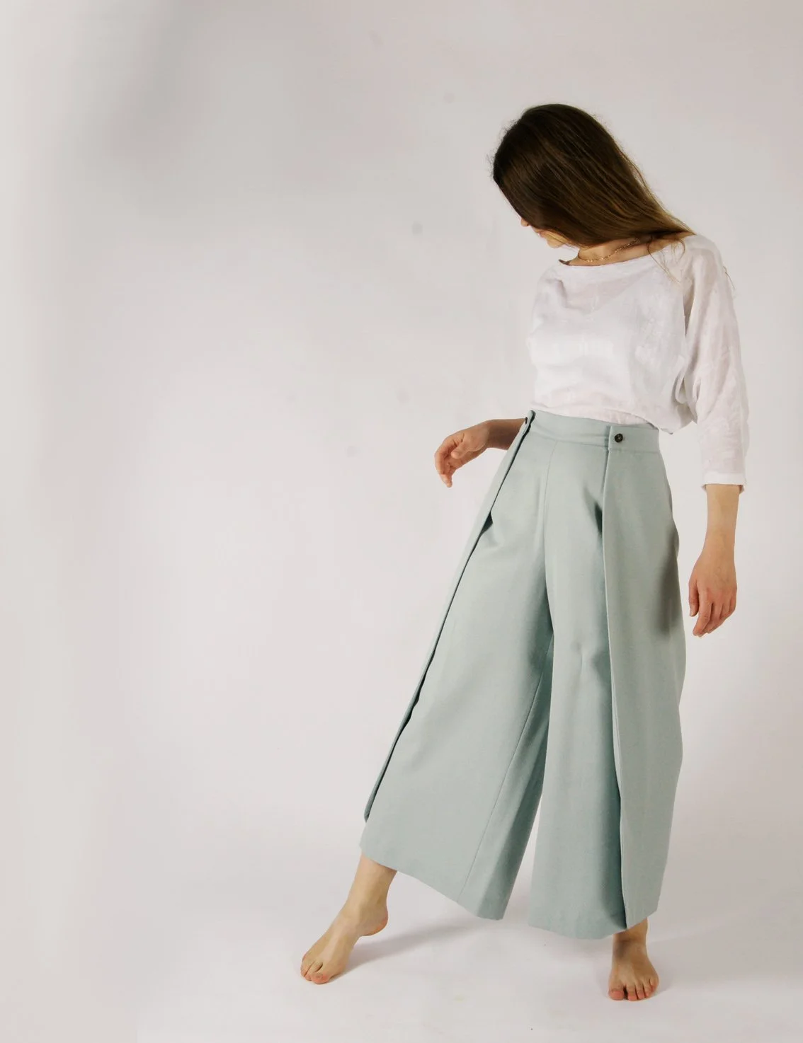 A woman in loose-fitting, wide-leg gray trousers with pleats, a white satin blouse with three-quarter sleeves, and no shoes, standing against a plain white background.