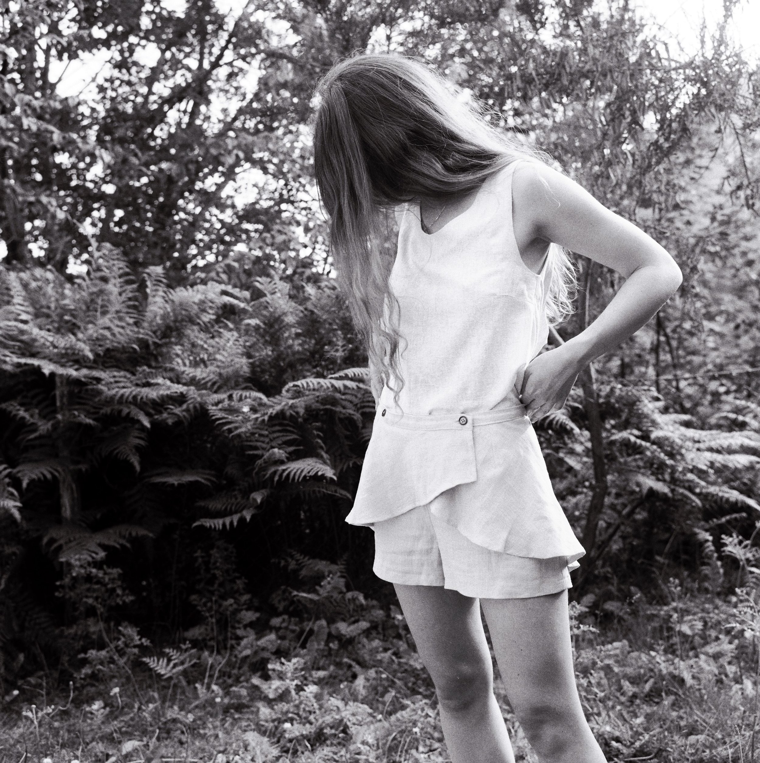 A woman with long hair wearing a sleeveless top and shorts in a forested area with ferns and trees.