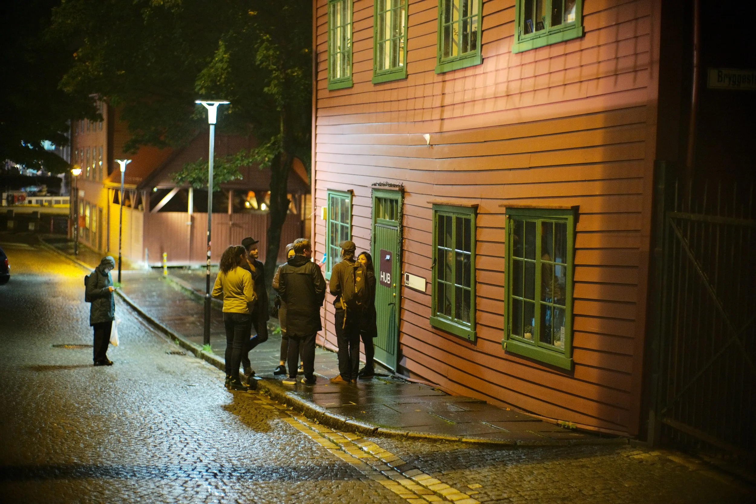 A group of people standing outside on a wet street at night, near a pink wooden building with green window frames. The scene is illuminated by streetlights.
