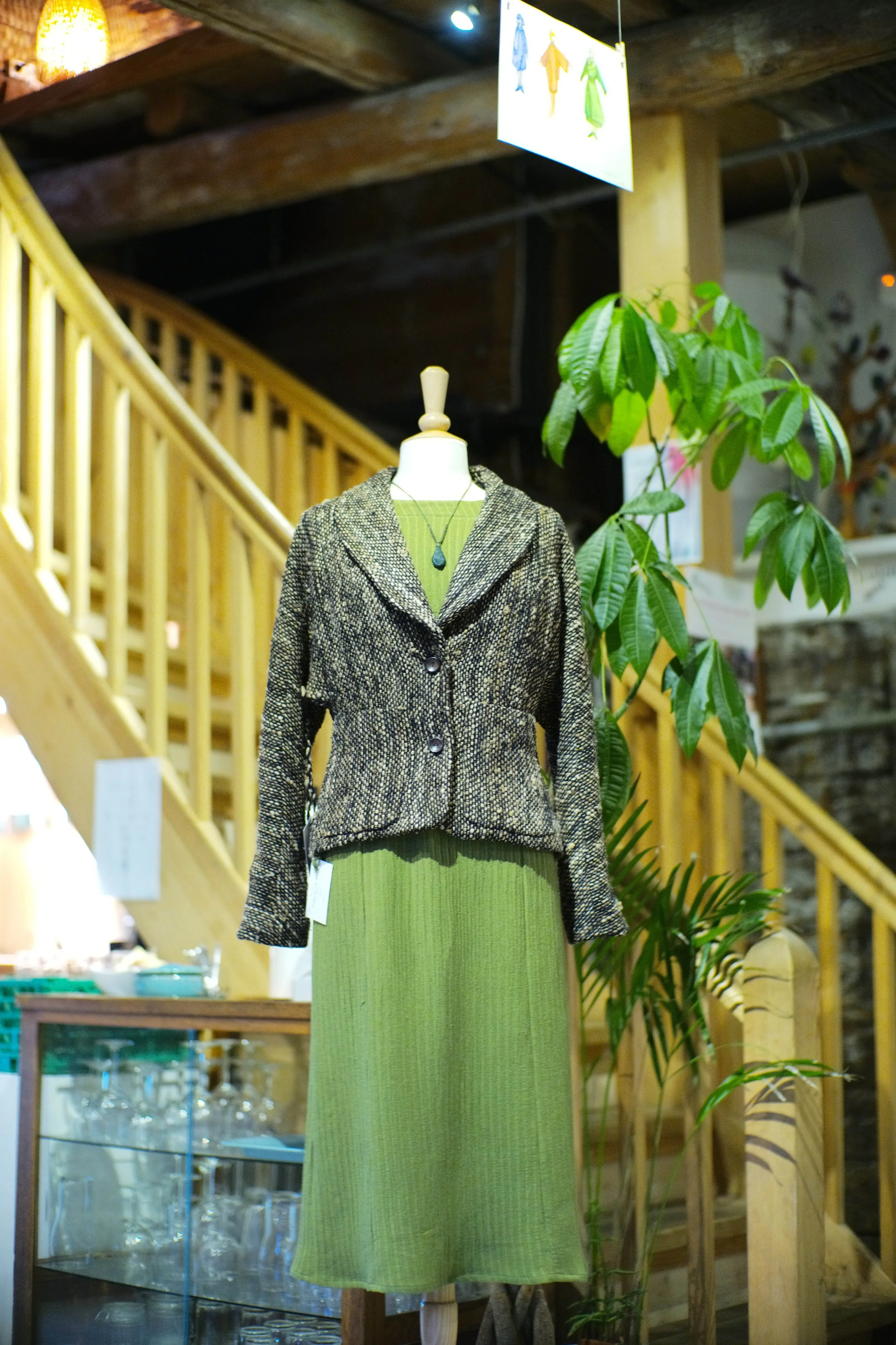 A mannequin dressed in a green ribbed dress and a patterned blazer, with a teardrop-shaped pendant necklace. The mannequin is positioned in a rustic wooden interior, with a staircase and leafy green plants nearby.