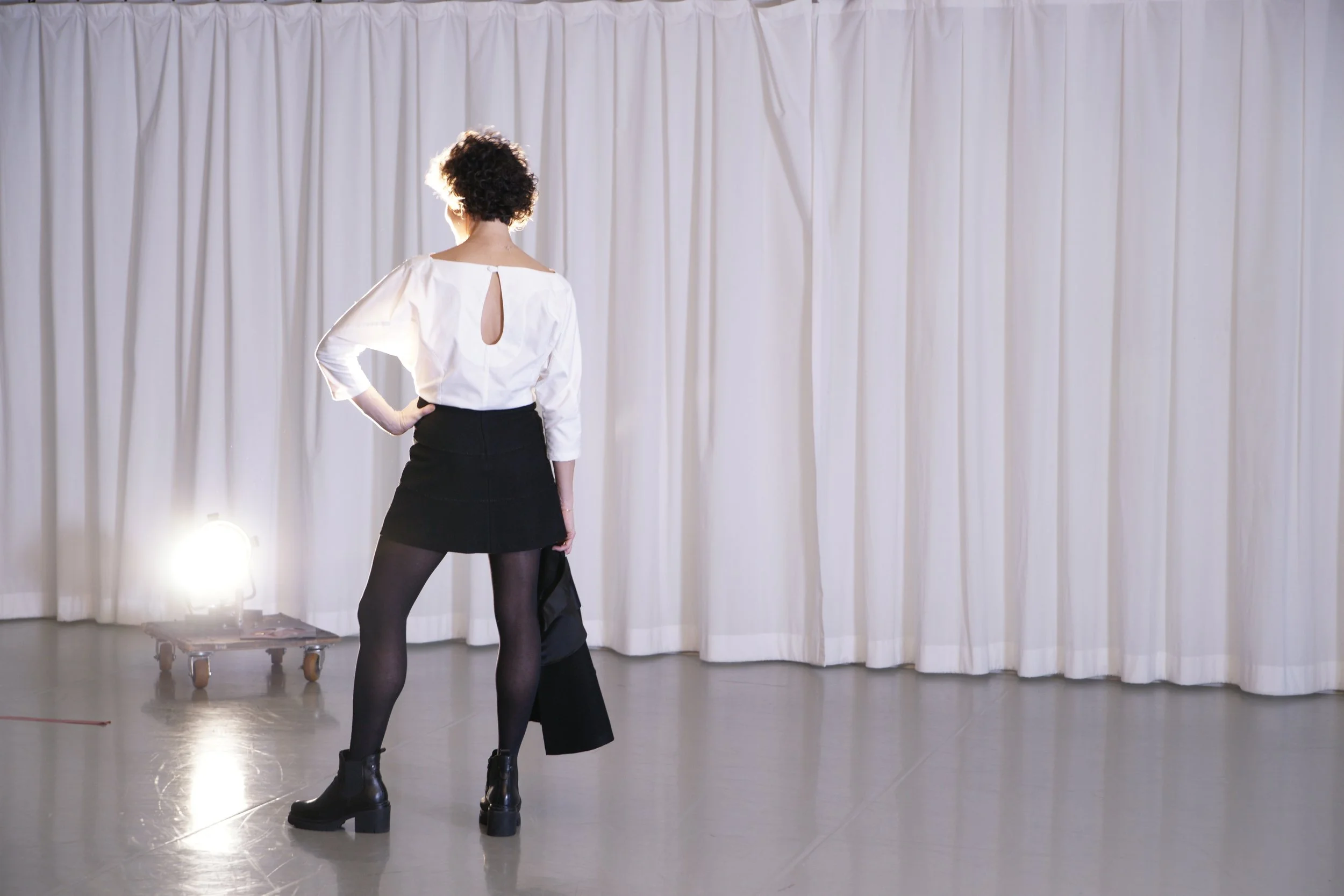 A woman with short curly hair wearing a white blouse with a keyhole back, black skirt, and black stockings standing with her hand on her hip in front of a white curtain, with stage lights and a cart with equipment nearby.