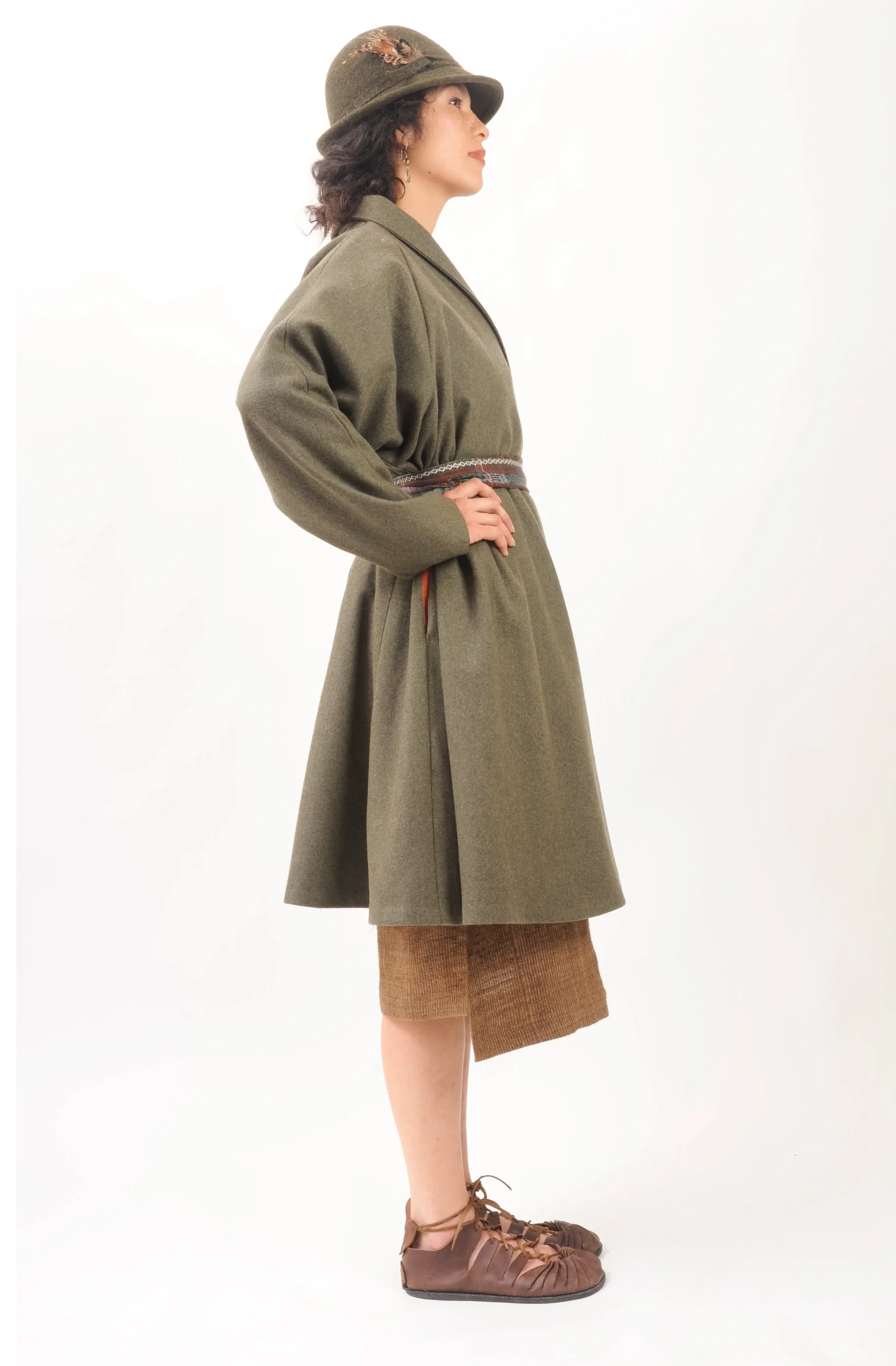 Side view of a woman in vintage inspired outfit, wearing a green coat, brown shorts, and tan lace-up shoes, with a hat decorated with feathers.