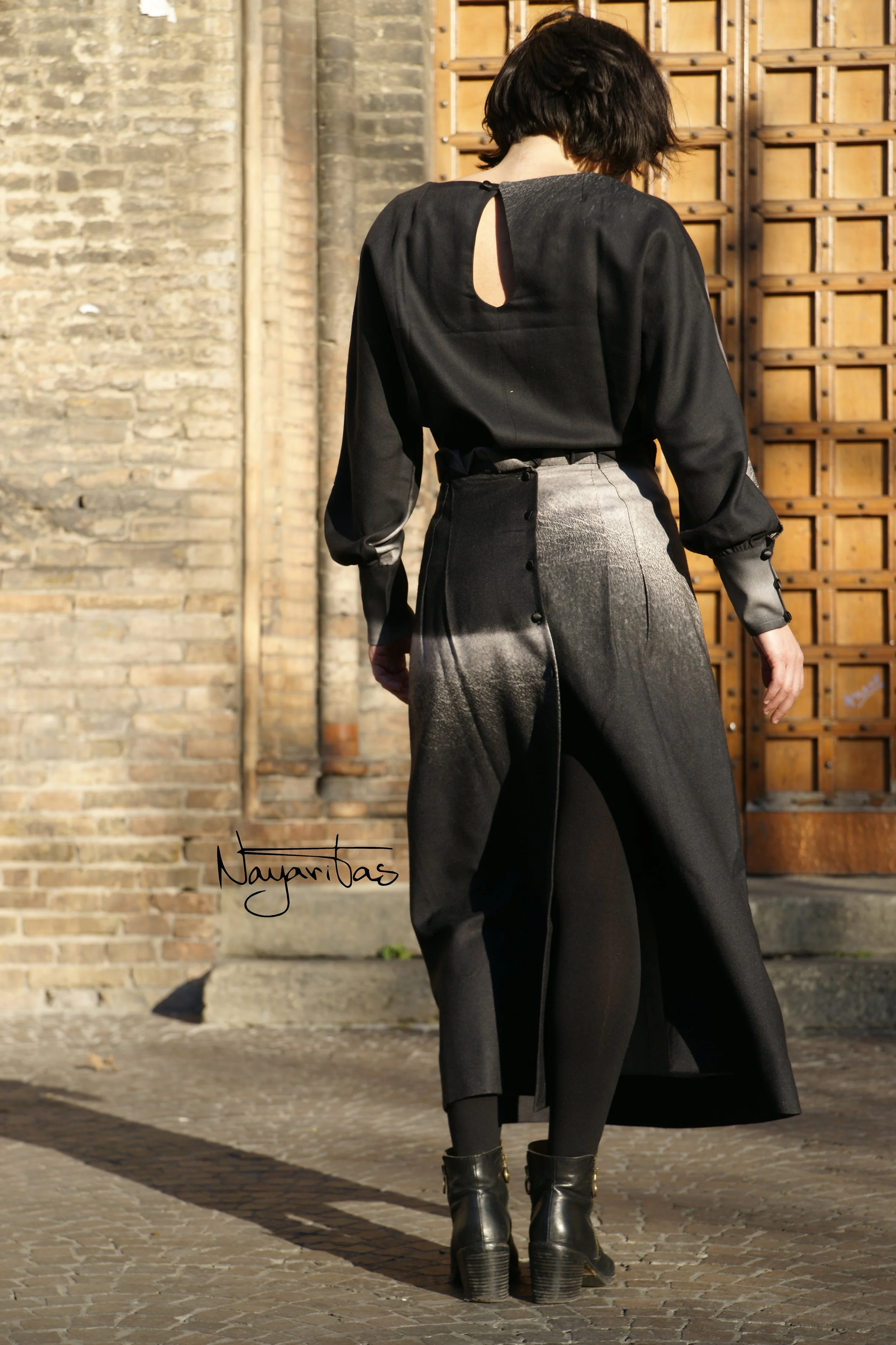 A woman with short dark hair wearing a black top with a keyhole back detail, long black skirt, and black ankle boots with heels, standing outdoors on a cobblestone street near a brick wall and a wooden gate.