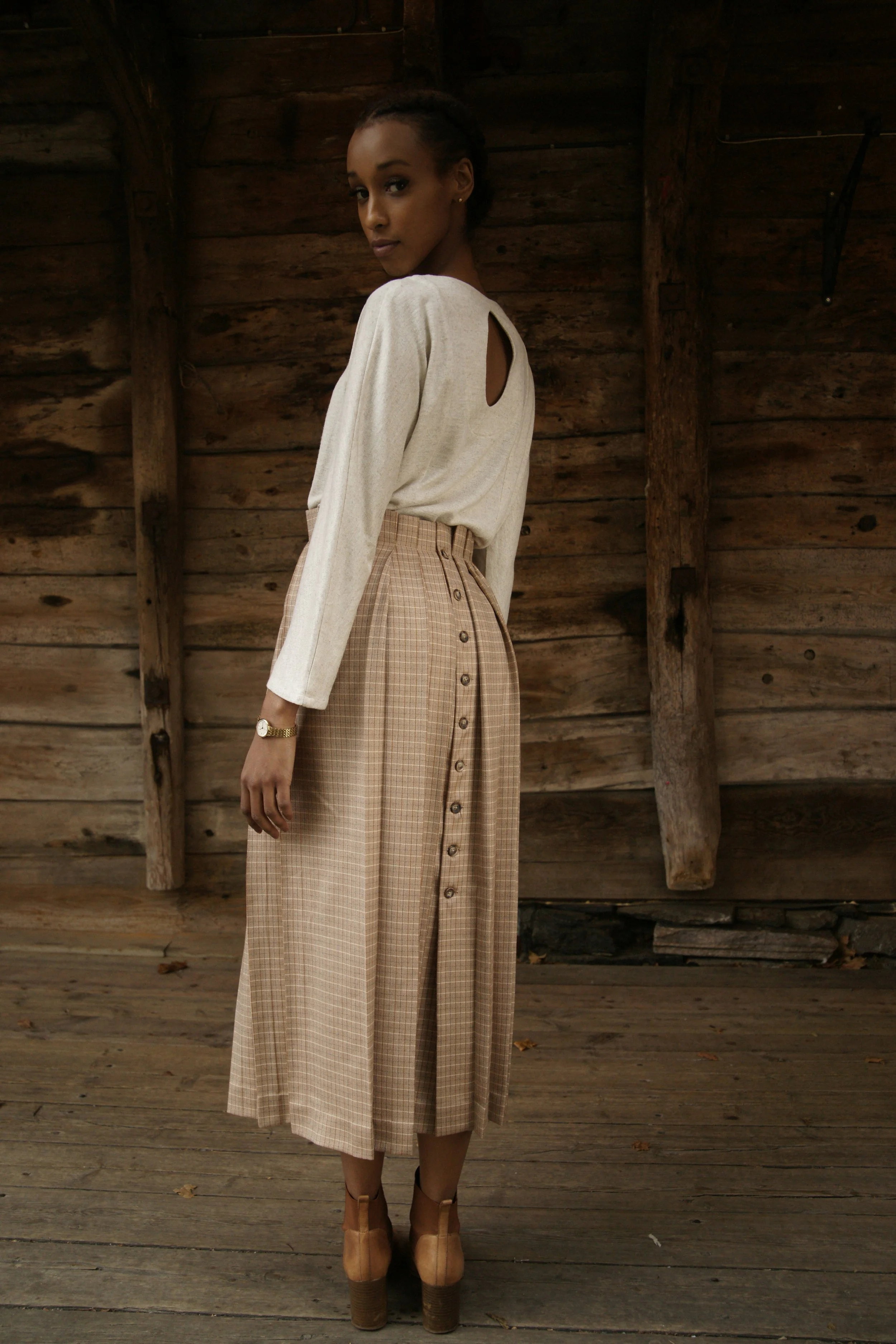 A woman standing against a wooden wall, wearing a beige long-sleeve top with a keyhole opening and a high-waisted plaid skirt with buttons down the front, paired with tan platform heels and a gold watch.