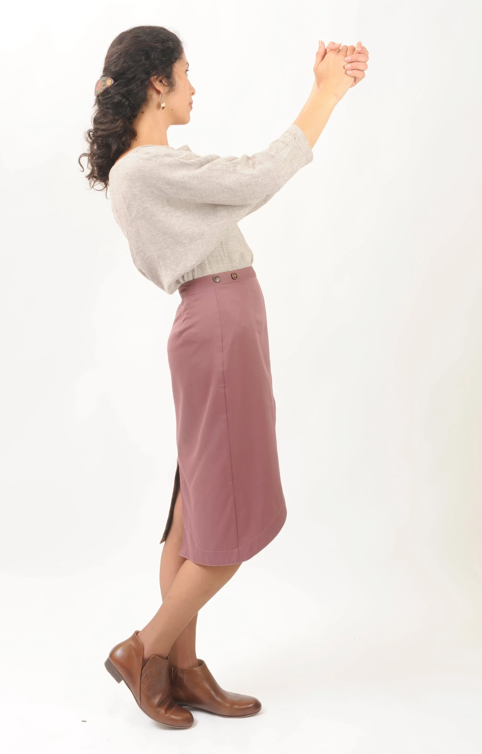 A woman standing sideways, wearing a beige top, a mauve skirt, and brown ankle boots, with her arms raised and hands clasped.