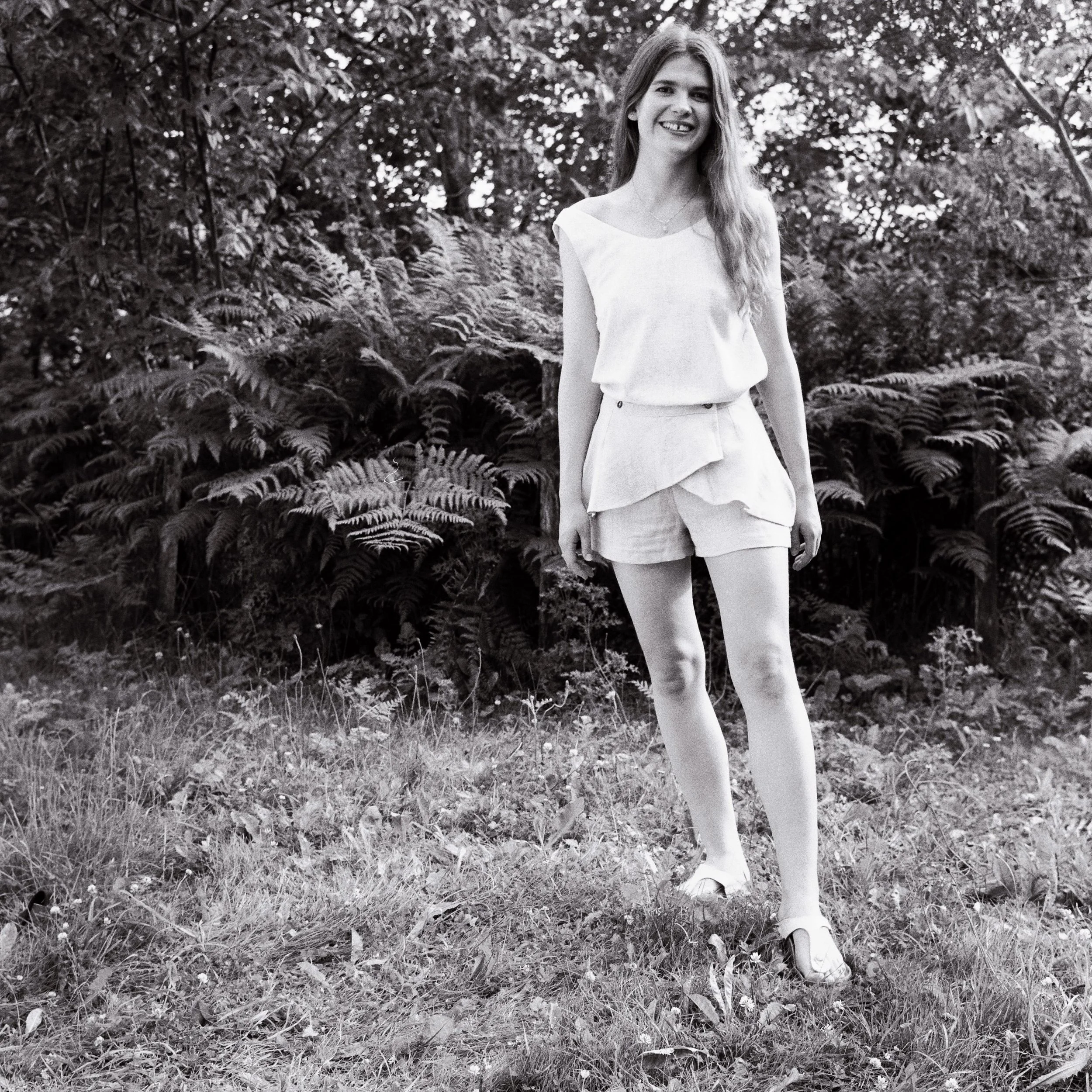 A woman with long hair smiling and standing outdoors among plants and trees, in black and white.