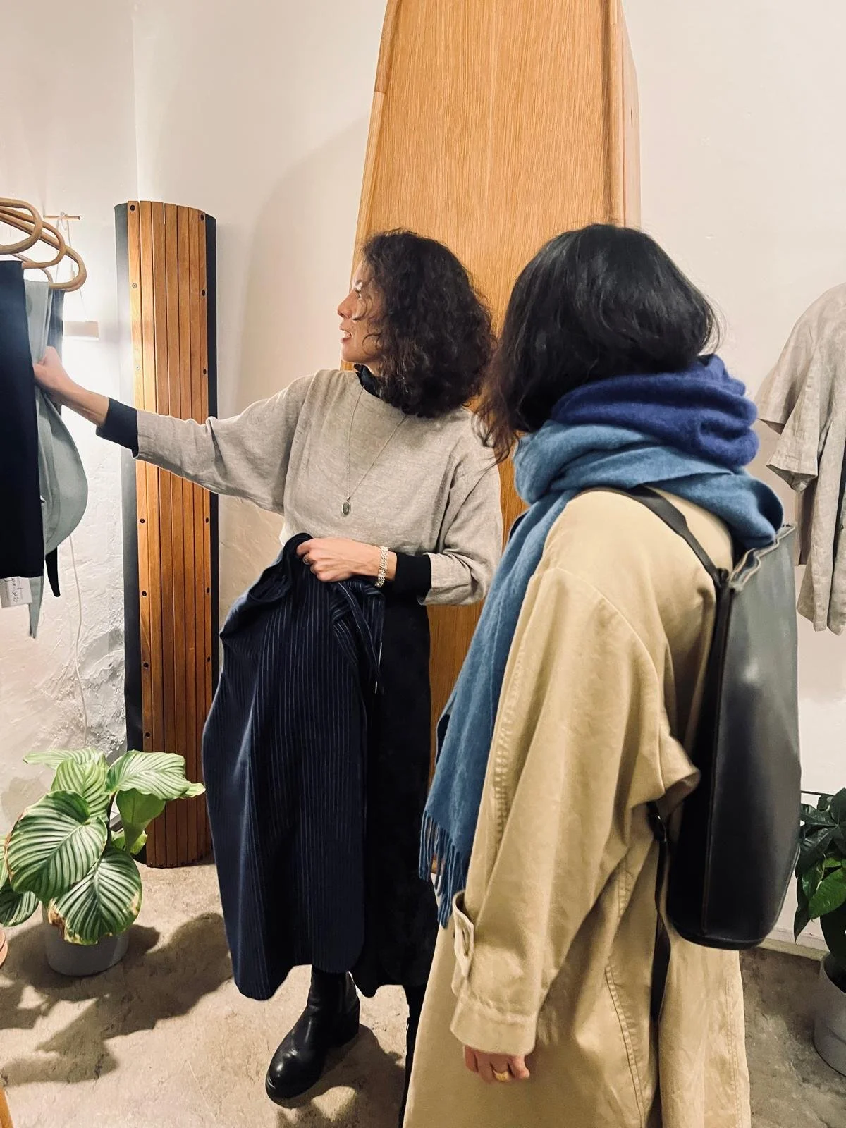 Two women shopping for clothes indoors, one reaching for a garment on a rack, the other holding a jacket. They are standing near a wooden wall and a potted plant.