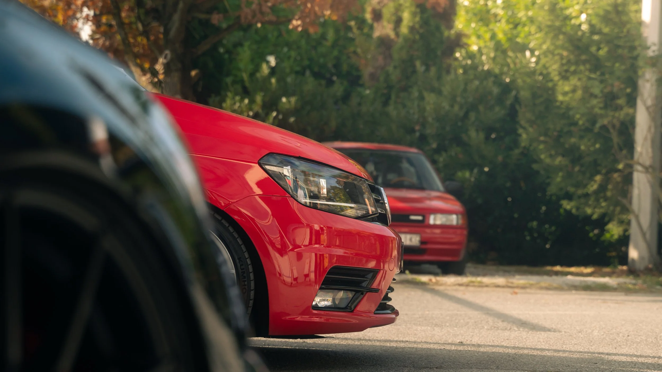 Close-up of the front of a red sports car parked on a tree-lined street, with a second red vehicle in the background.