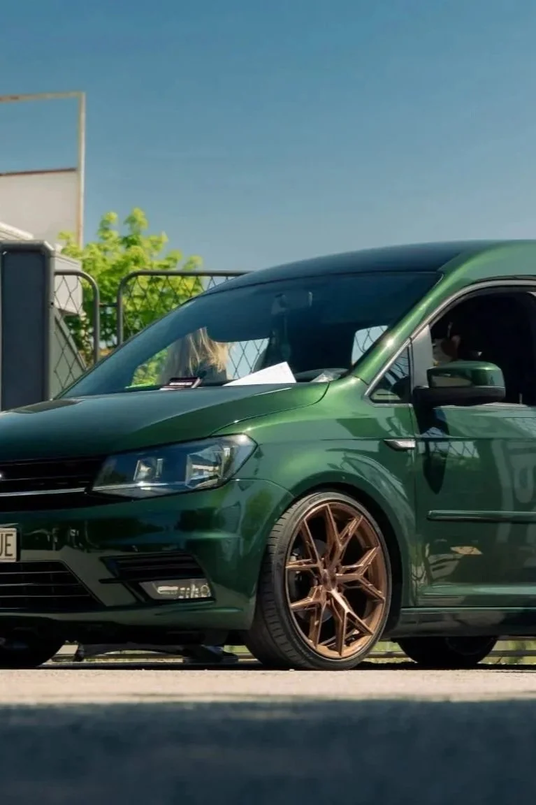 Green hatchback car with bronze-colored wheels parked outdoors under a blue sky, with a person sitting in the driver’s seat.