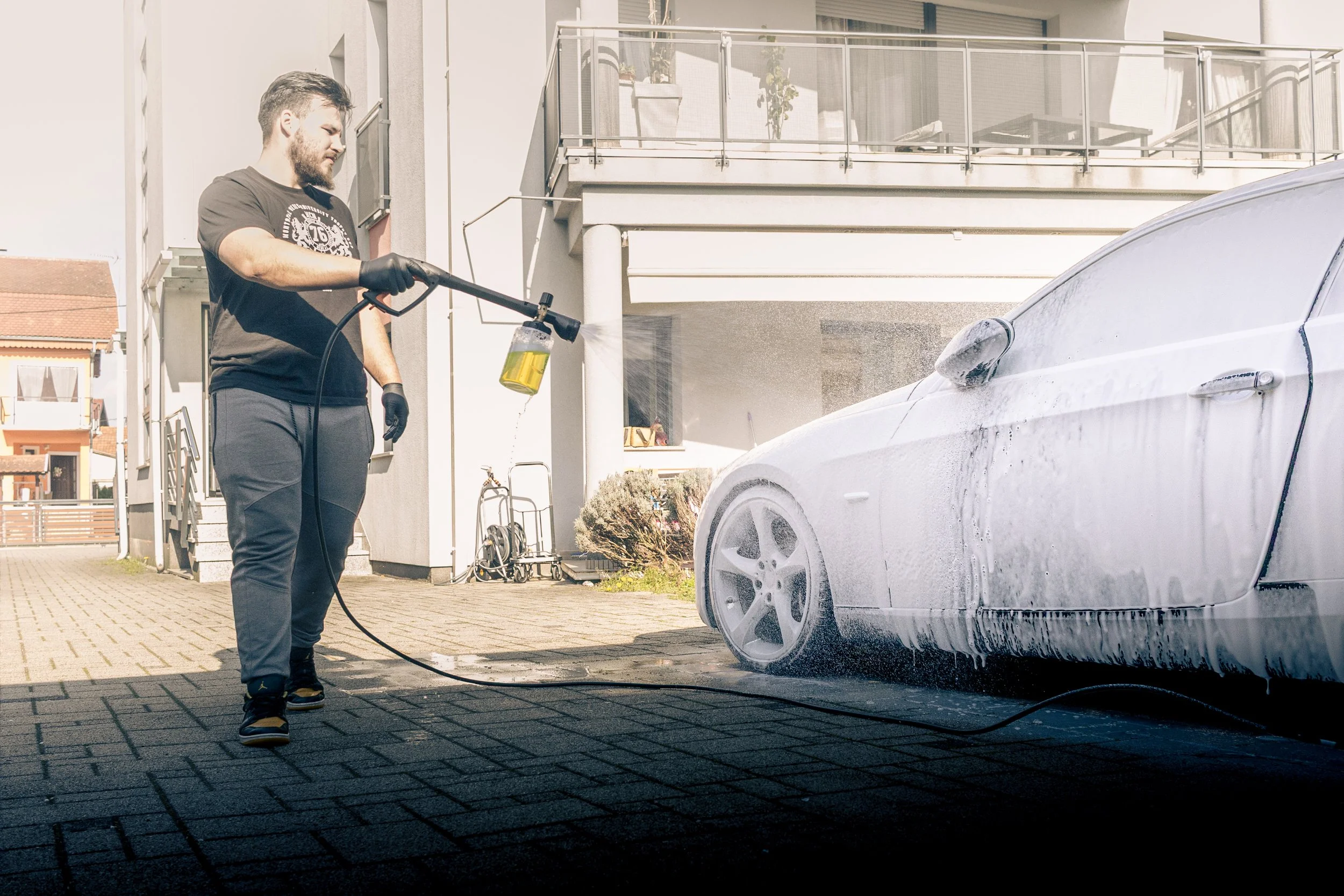 A man is holding a pressure washer and spraying foam on a white car parked outside a residential building.