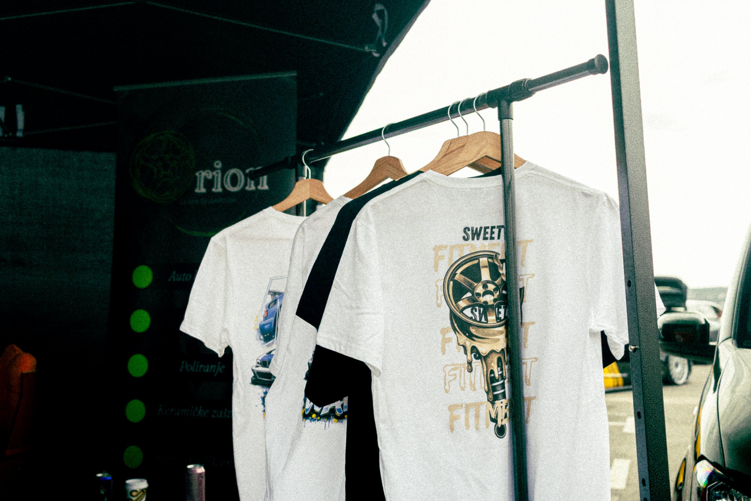 Clothing rack displaying t-shirts with graphic designs, including one with an ice cream cone and the words "FOMO" and "FOMO FIT" in a parking lot.