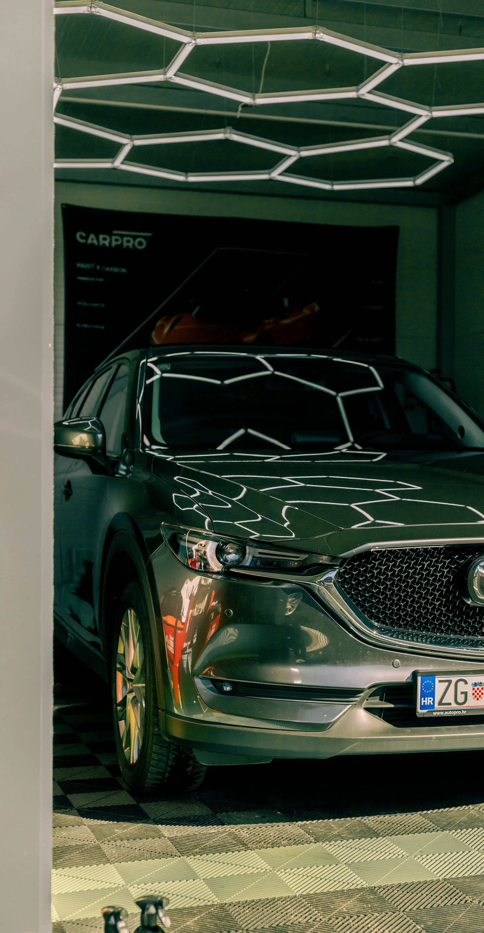 Silver SUV inside a car showroom with geometric ceiling lights reflected on its surface. The front grille and headlights are visible, and part of a Croatian license plate can be seen.