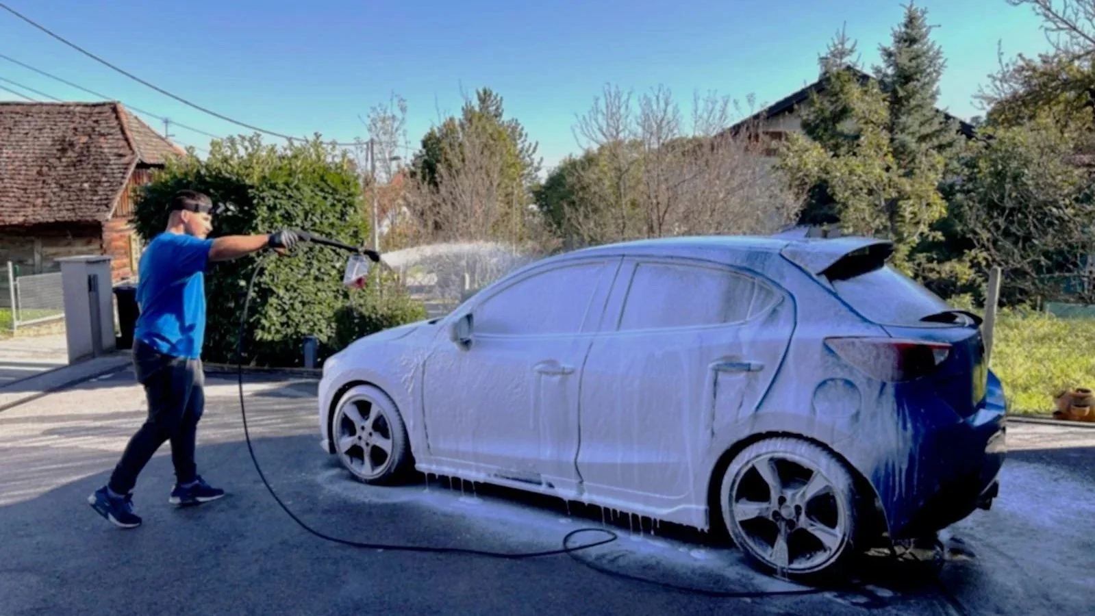 A person wearing a blue shirt, black pants, and gloves is washing a blue car with a hose, creating a lot of soap suds on the vehicle in an outdoor driveway.