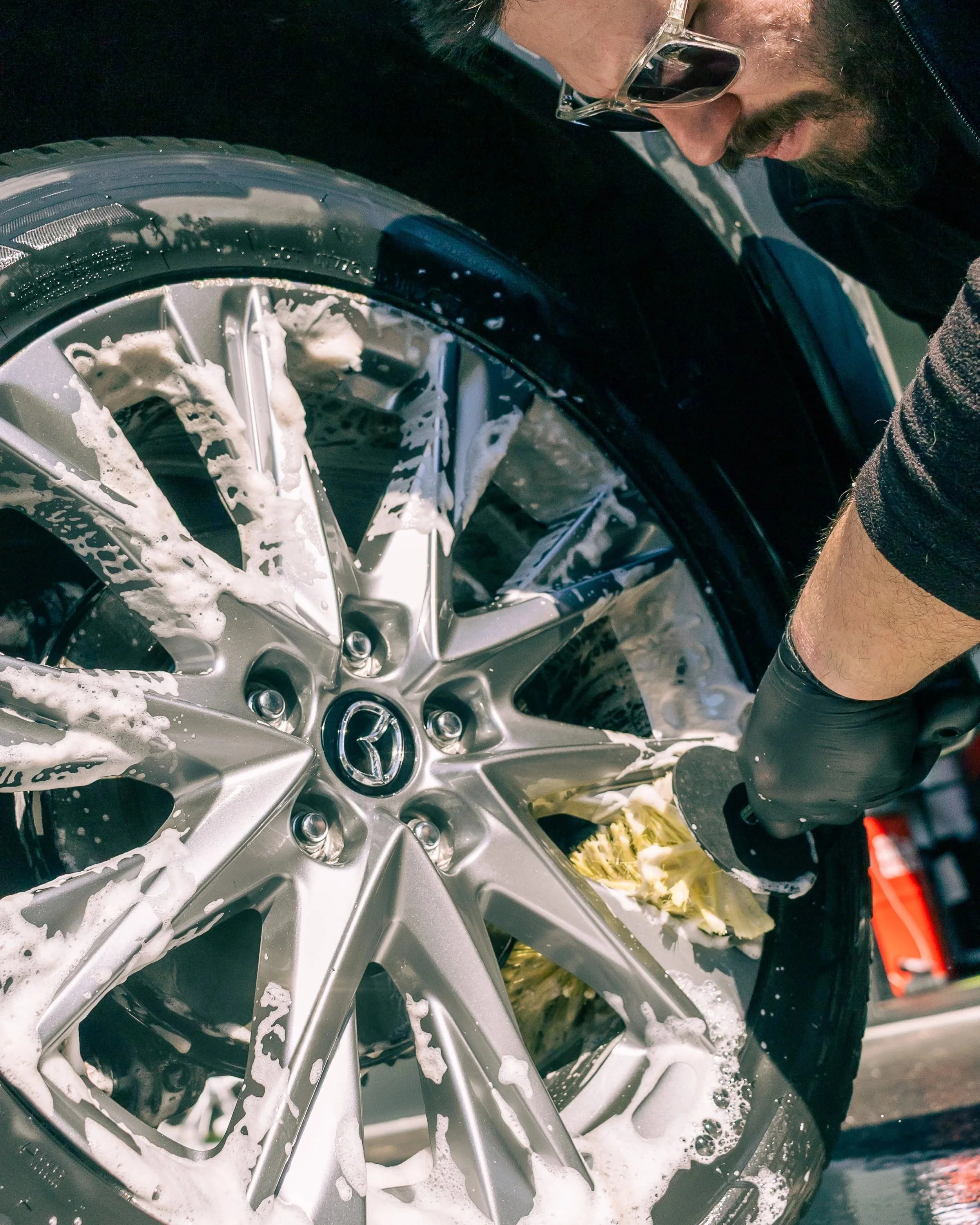 A person cleaning a silver Honda car wheel with soap, foam, and a brush.