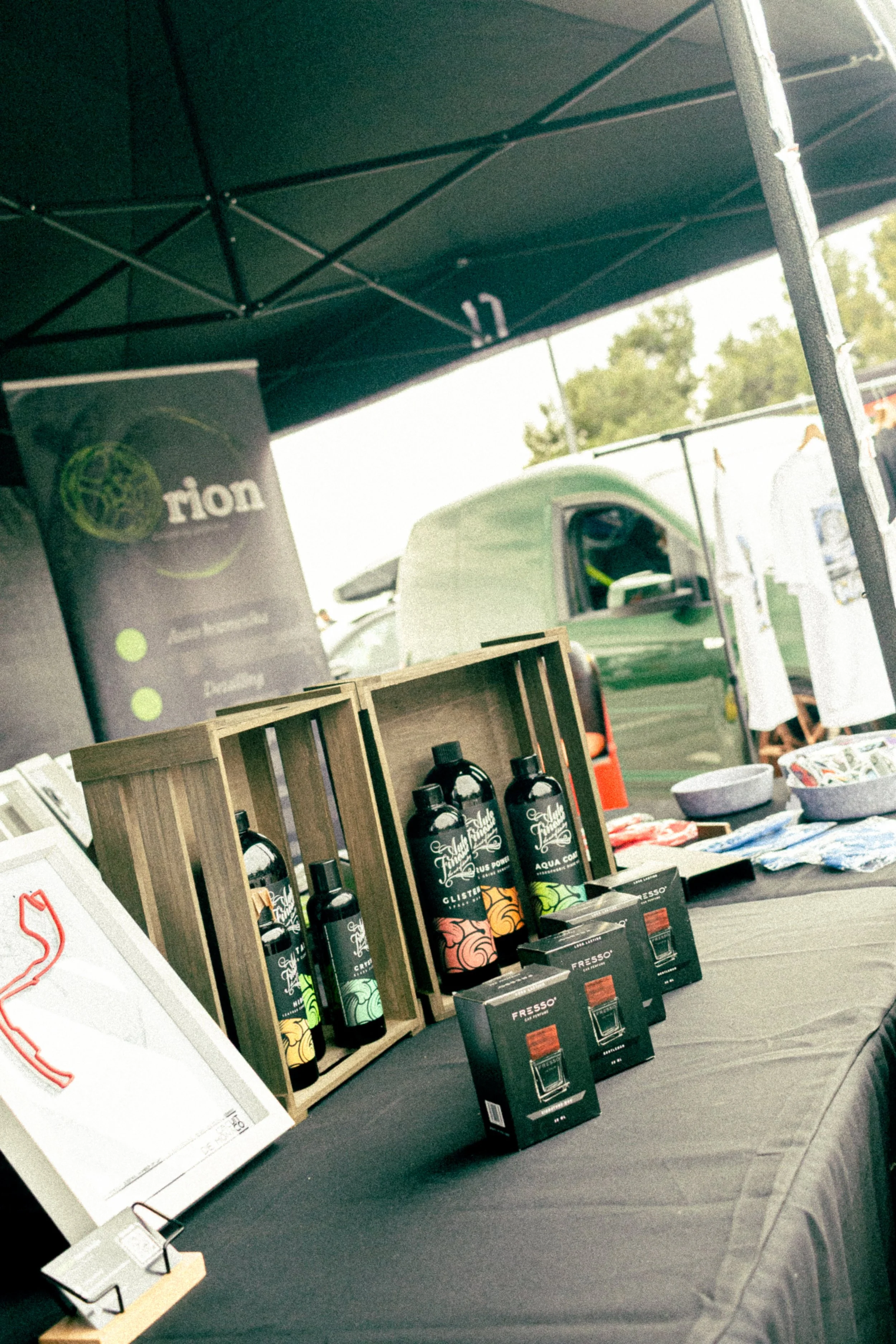 Table display at an outdoor event with bottles, boxes, and promotional materials, including a banner with green and yellow accents and a black tent overhead.