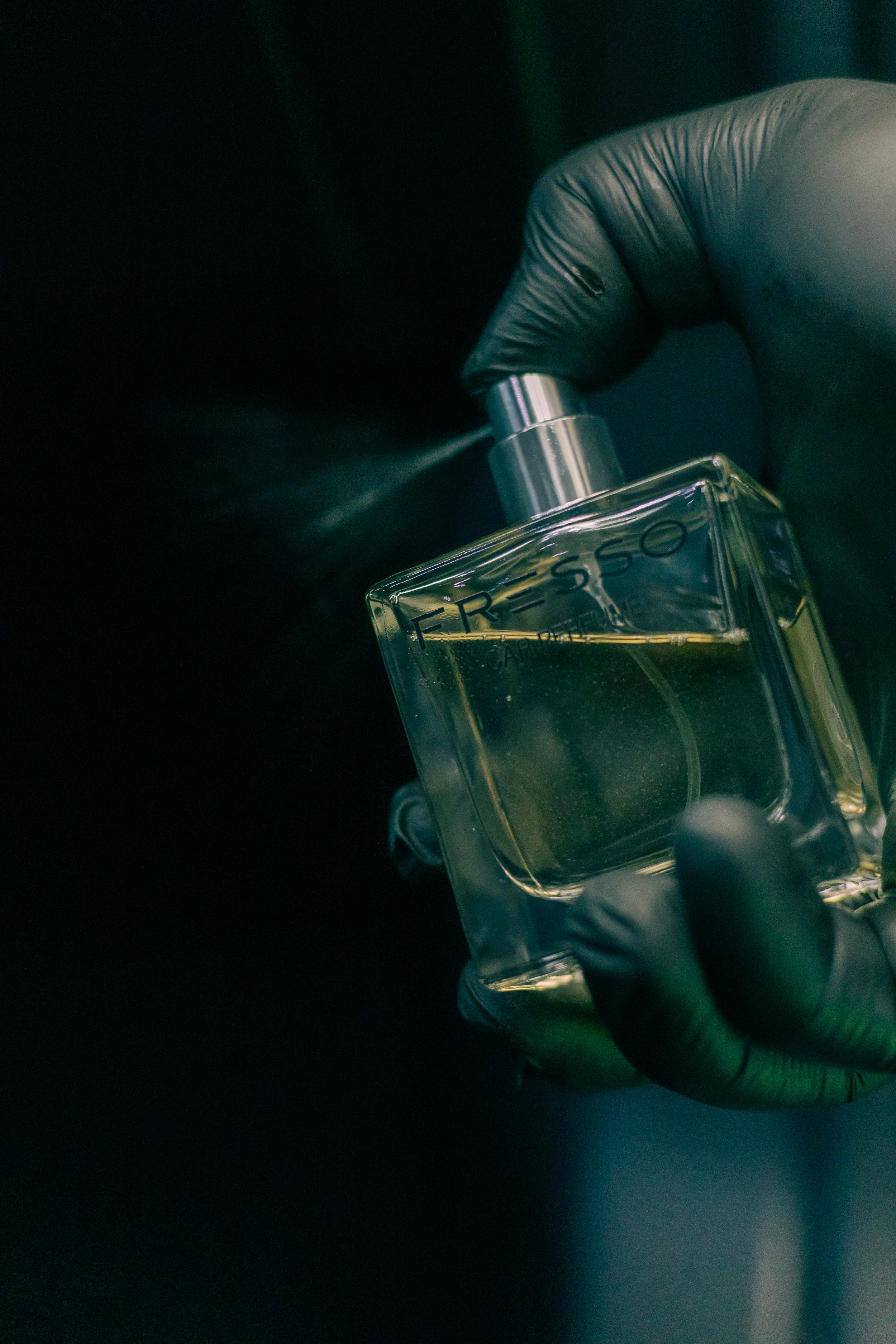 A hand wearing a black glove holding a transparent perfume bottle labeled 'Resso.' The person is spraying perfume onto their wrist.