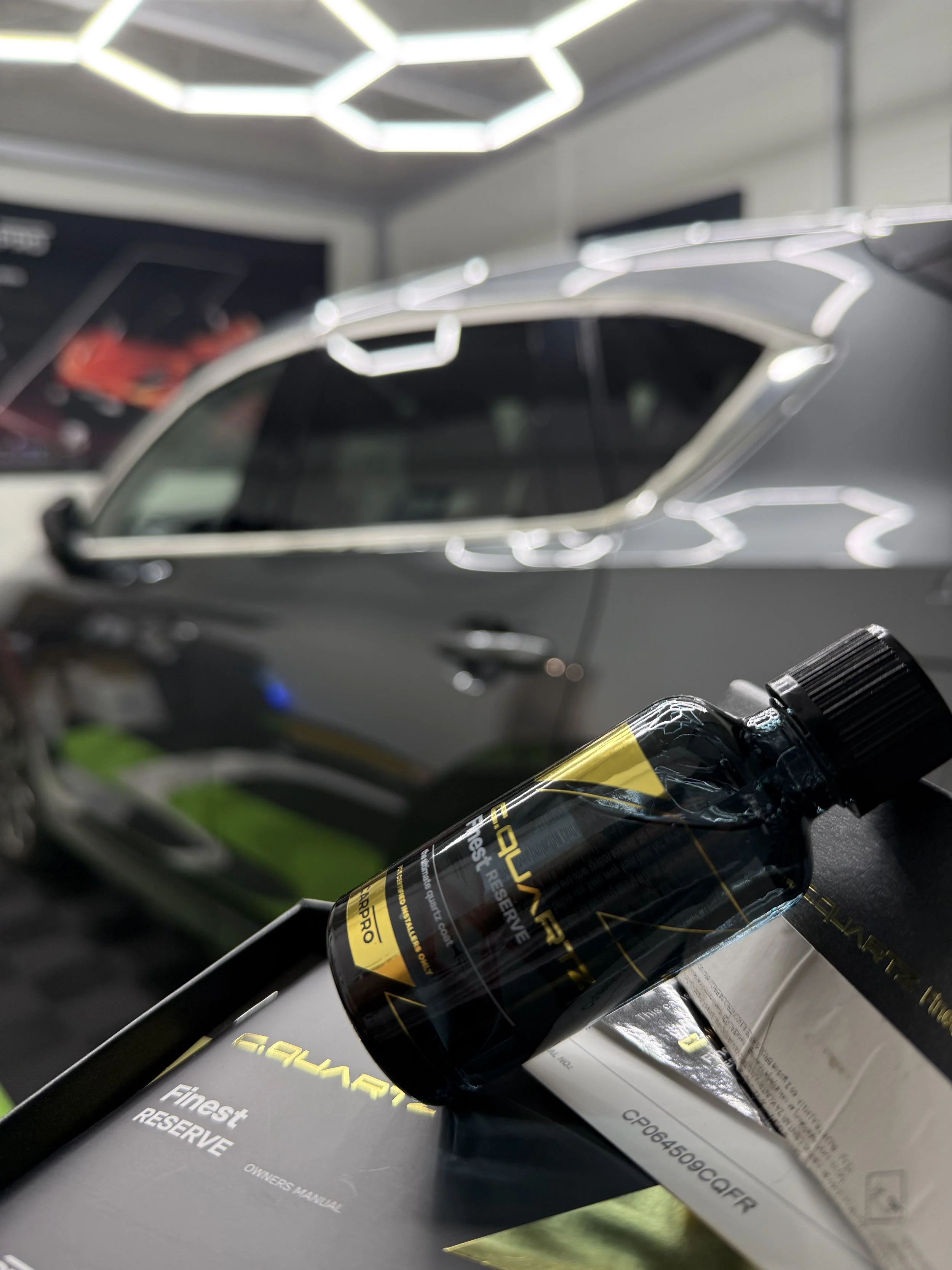A black bottle of Meguiar's Hyper Dressing Restores and Protects appears in the foreground. In the background, there's a partially visible gray car with a shiny finish and a modern headlight design. The car is on display at a showroom or workshop with bright LED lighting on the ceiling.