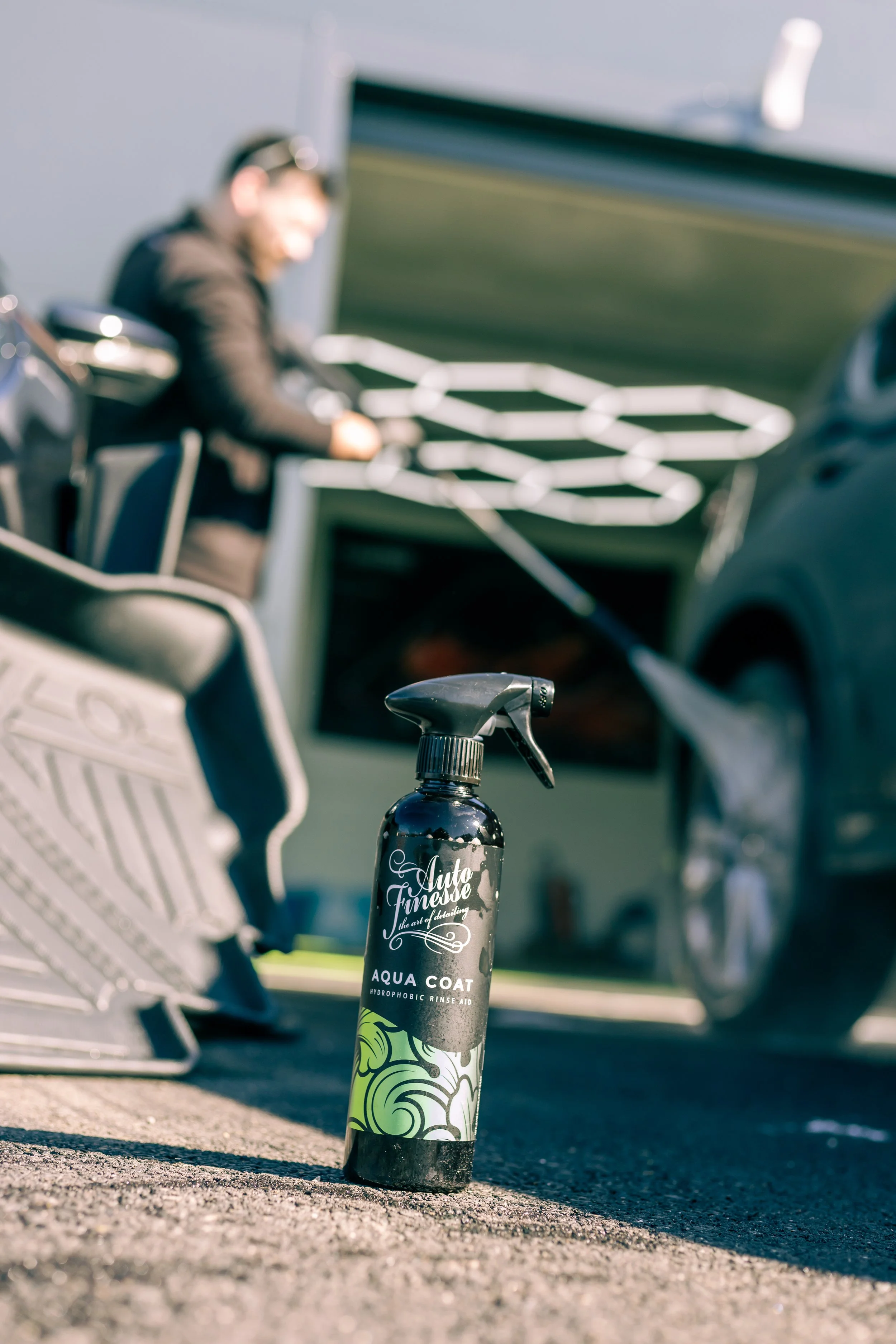 A spray bottle labeled 'Aqua Coat' standing on concrete ground at a car detailing or cleaning station, with blurred person and car in the background.