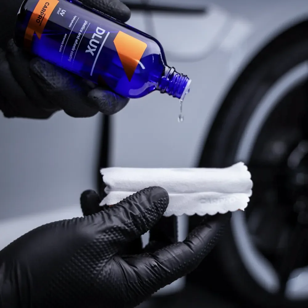 A person wearing black gloves applying CARPRO DLUX ceramic coating to a white sponge applicator, with a blue bottle of the product in hand and a car tire in the background.
