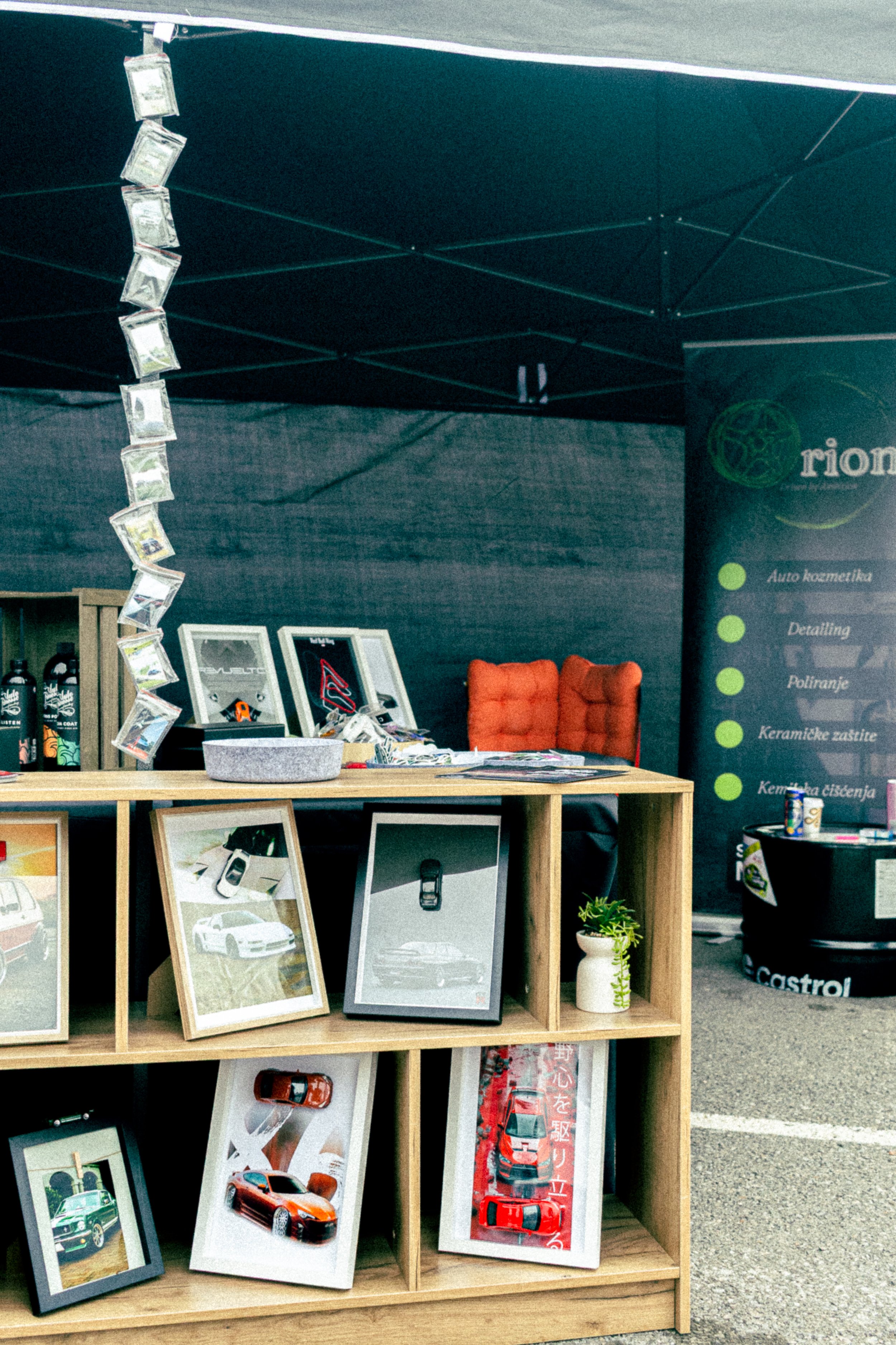 Display of framed car photographs on a wooden shelf at an outdoor event, with a black tent in the background.