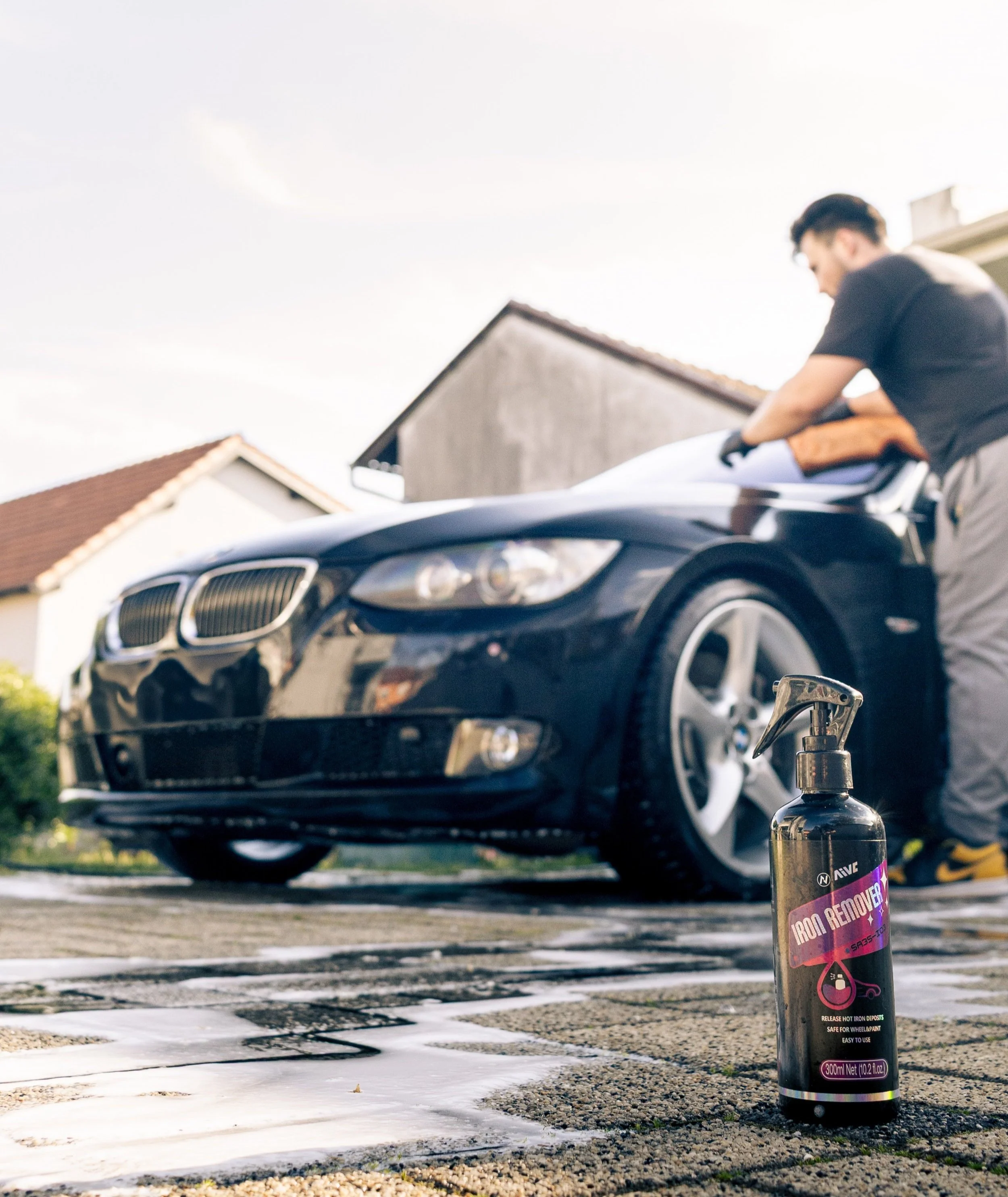 A man cleaning a black BMW car outside, with a bottle of iron remover on the ground in the foreground.