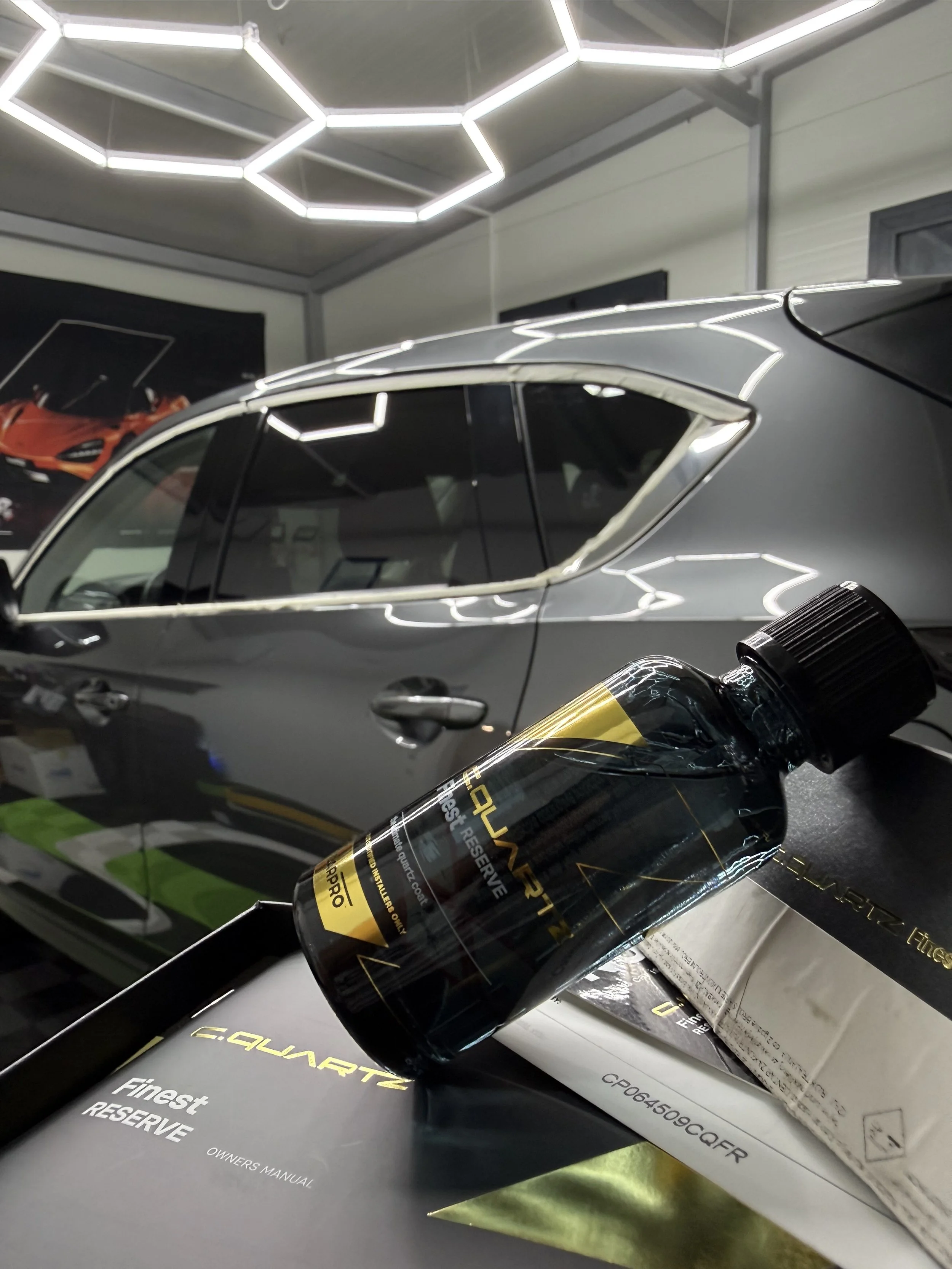Close-up of a black bottle labeled 'HYPERFINE Reserve' resting against a car and a box, with bright overhead geometric lighting reflections.