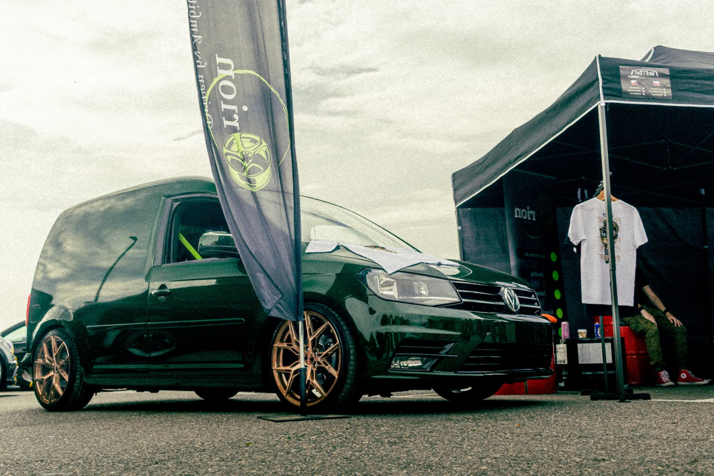 A black Volkswagen car with gold rims parked at an outdoor event booth, with promotional flags, a tent, and a person sitting to the right in the background under cloudy skies.