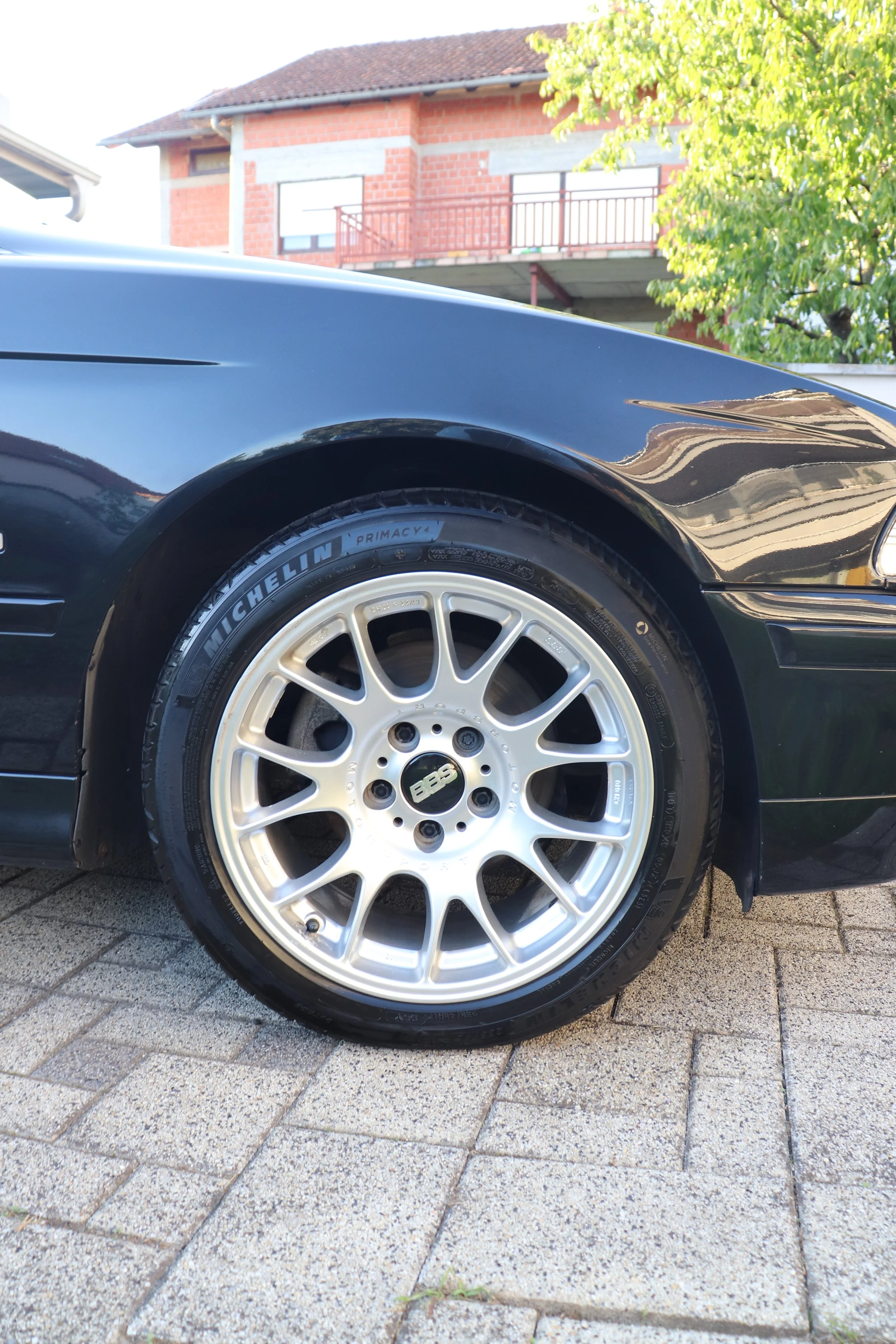 Close-up of a car's front wheel with a BBS alloy rim and Michelin Primacy 4 tire, parked on a paved driveway. Background includes a brick building and trees.