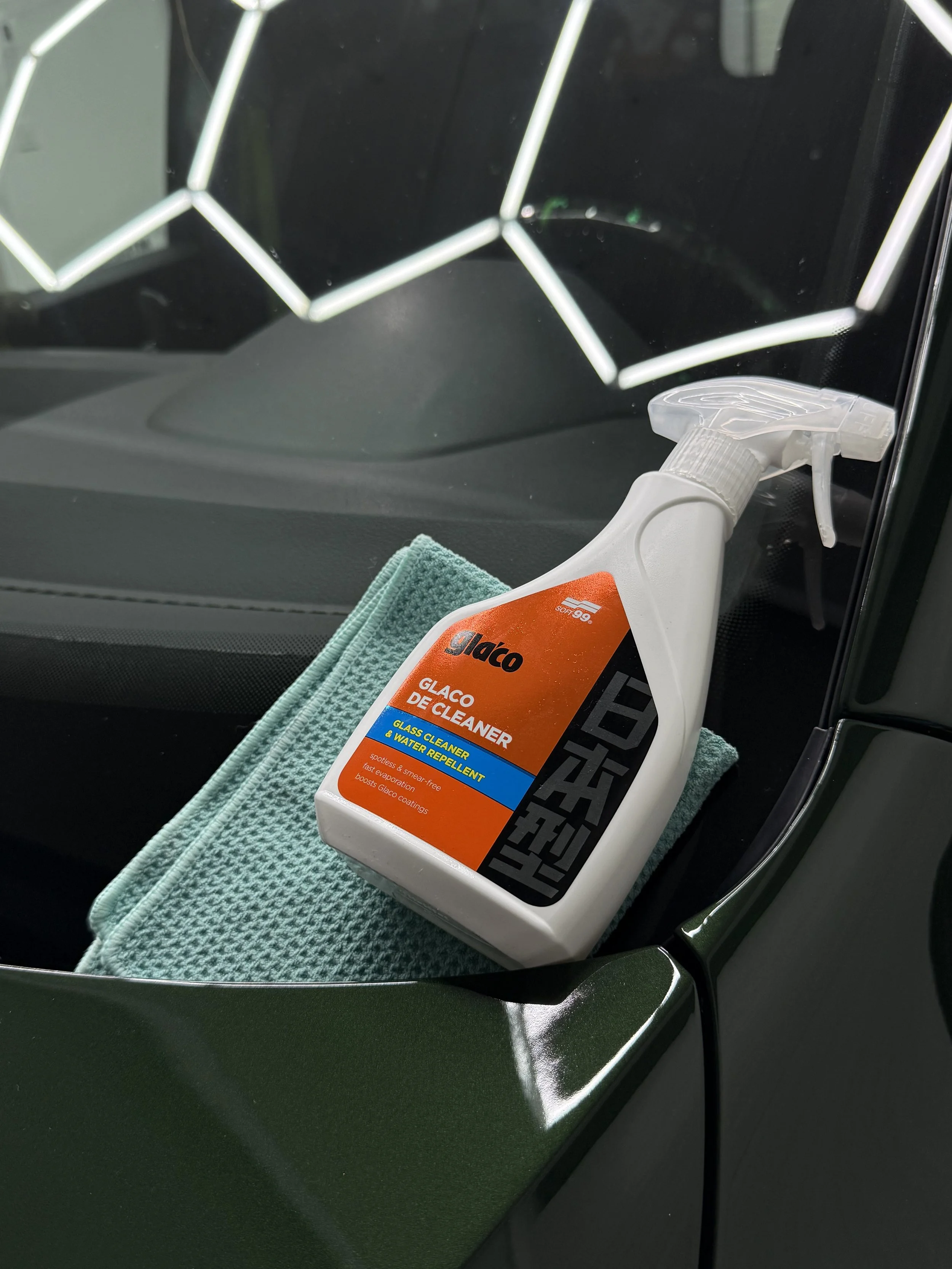 A green car dashboard with a microfiber cloth and a white spray bottle of glass cleaner resting on top.