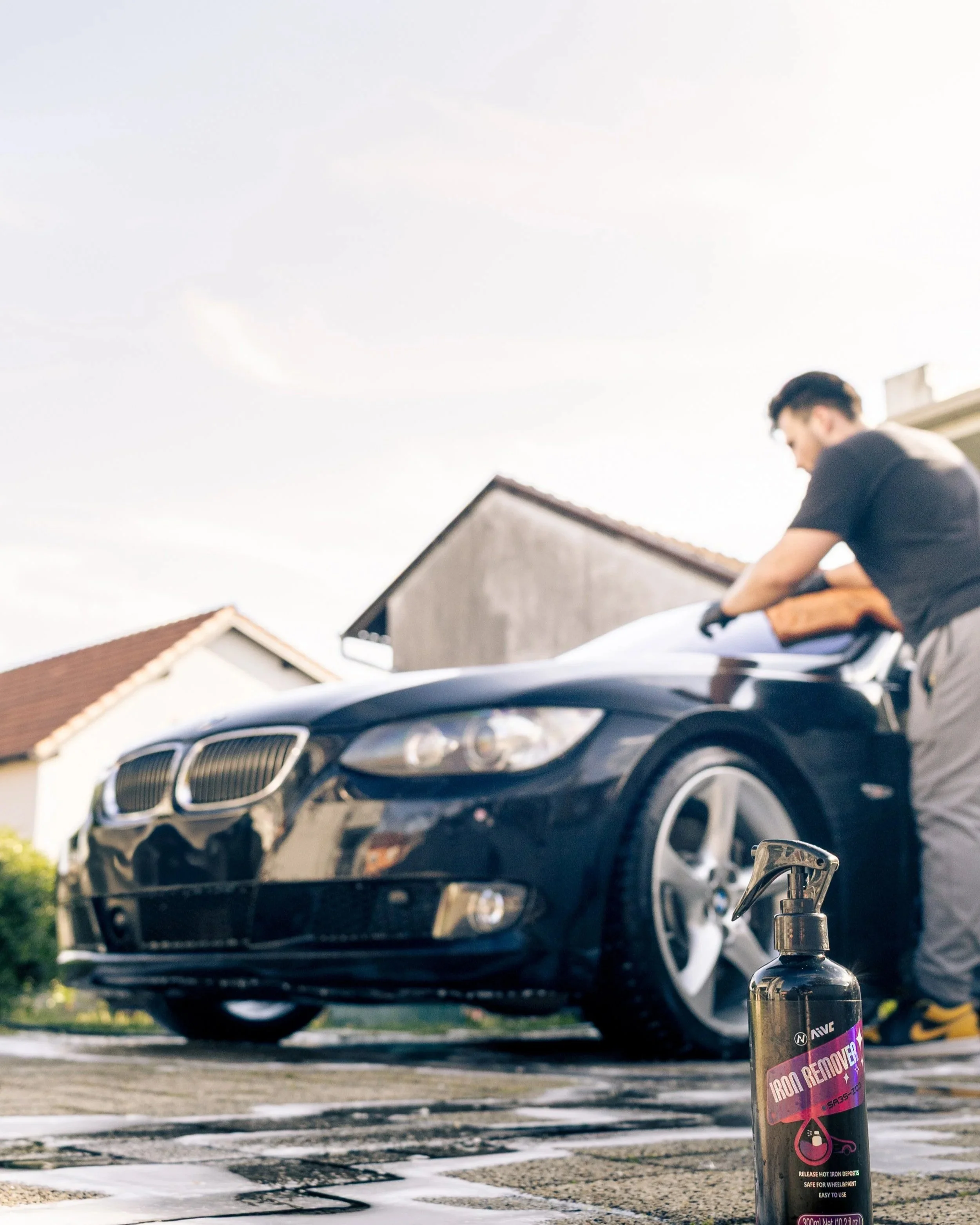 Car parked on driveway with a bottle of iron remover spray in the foreground and a man cleaning the car's hood in the background.