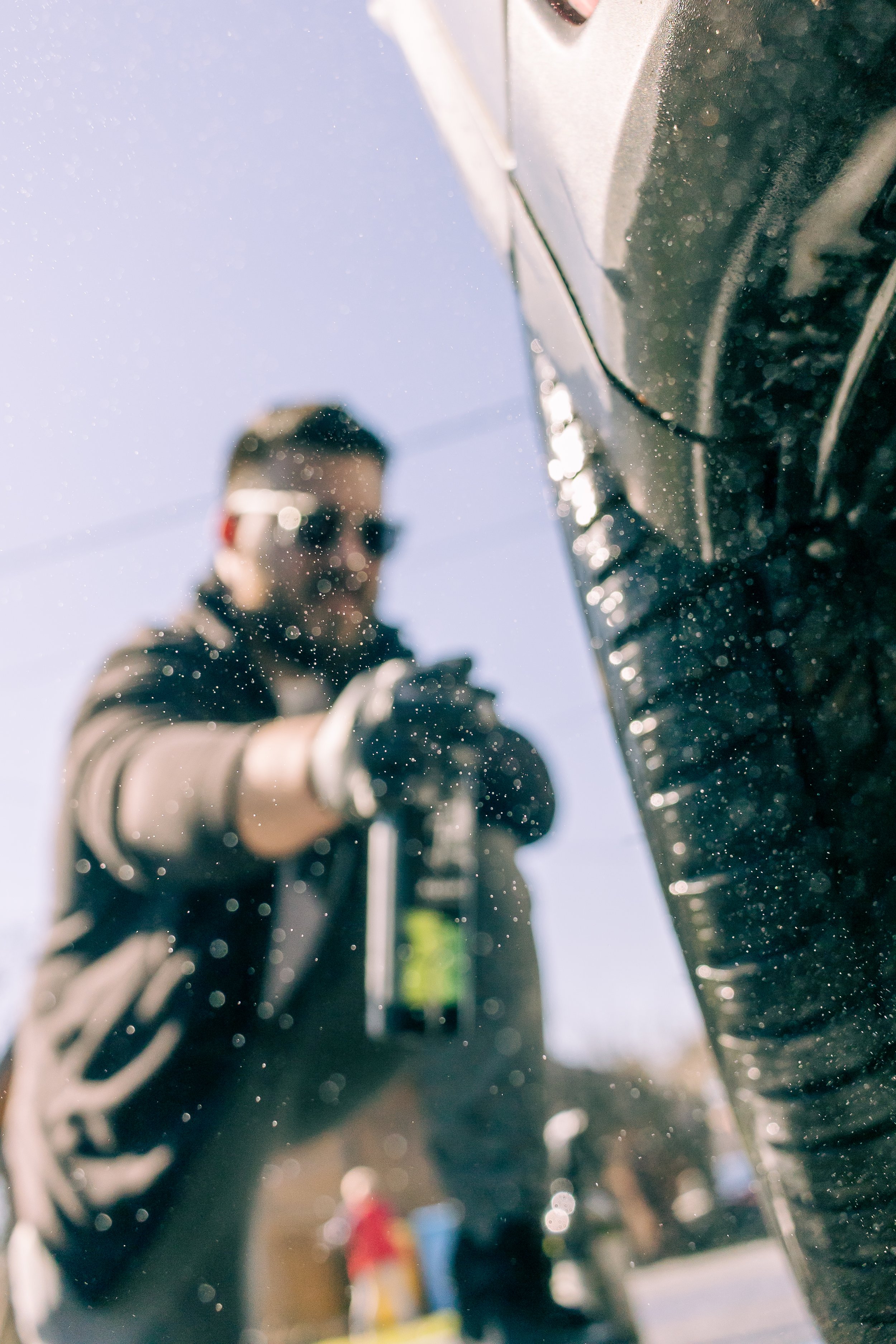 Close-up of a man in sunglasses taking a photo with a camera, reflected in a shiny surface with water droplets, with blurred background.