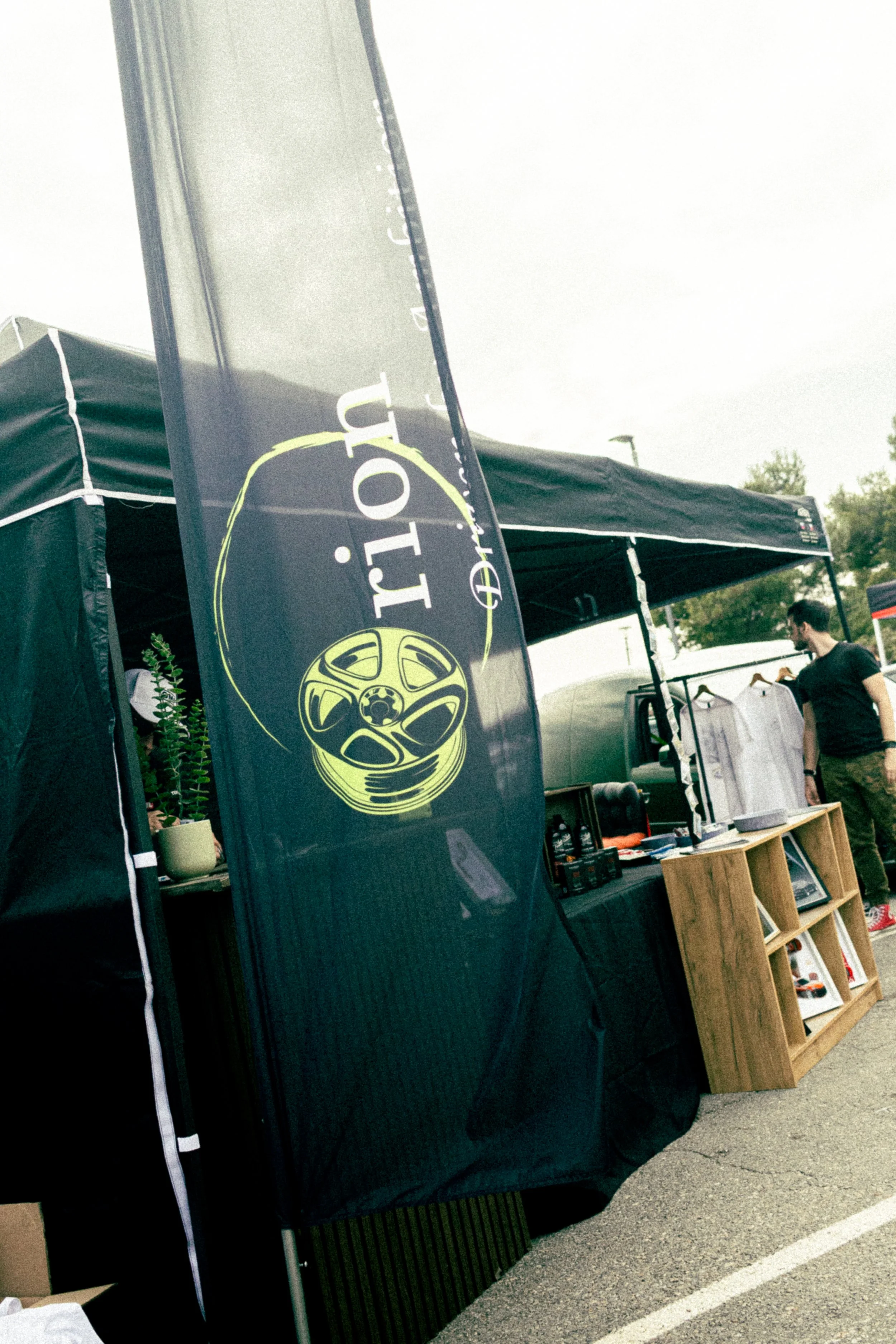 An outdoor stall with a black booth, a tall flag with a wheel design and the words 'lot' on it, and a wooden display shelf with framed pictures and shoes, with a person browsing items in the background.
