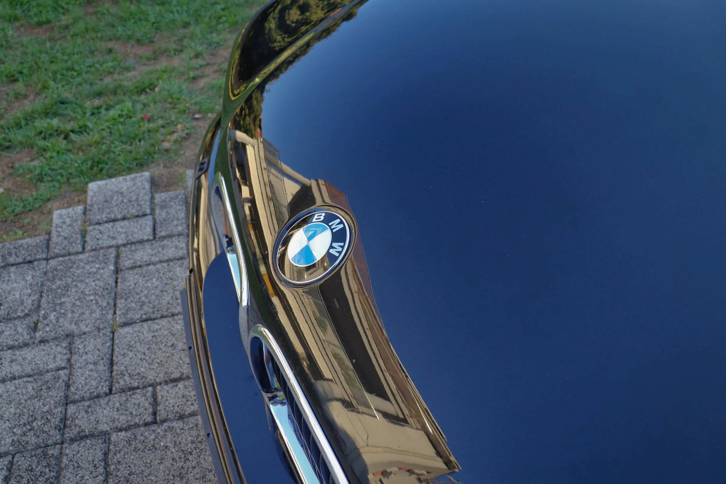 Close-up view of a shiny black BMW car hood with the BMW logo in the center, reflecting the surrounding environment, including a church steeple and trees, with a background of grass and stone paving.