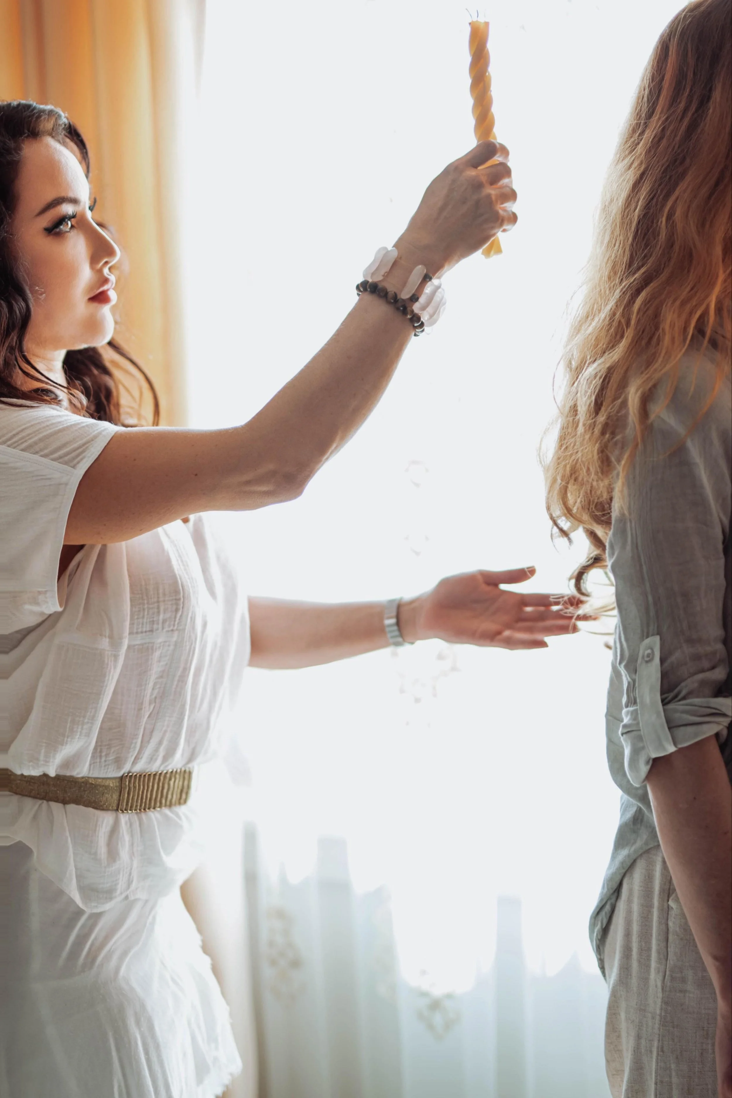 A woman with dark red, curly hair wearing a white shirt and beige belt is holding a twisted yellow candle in front of another woman with long blonde hair wearing a gray top, both standing indoors near a window with bright sunlight.