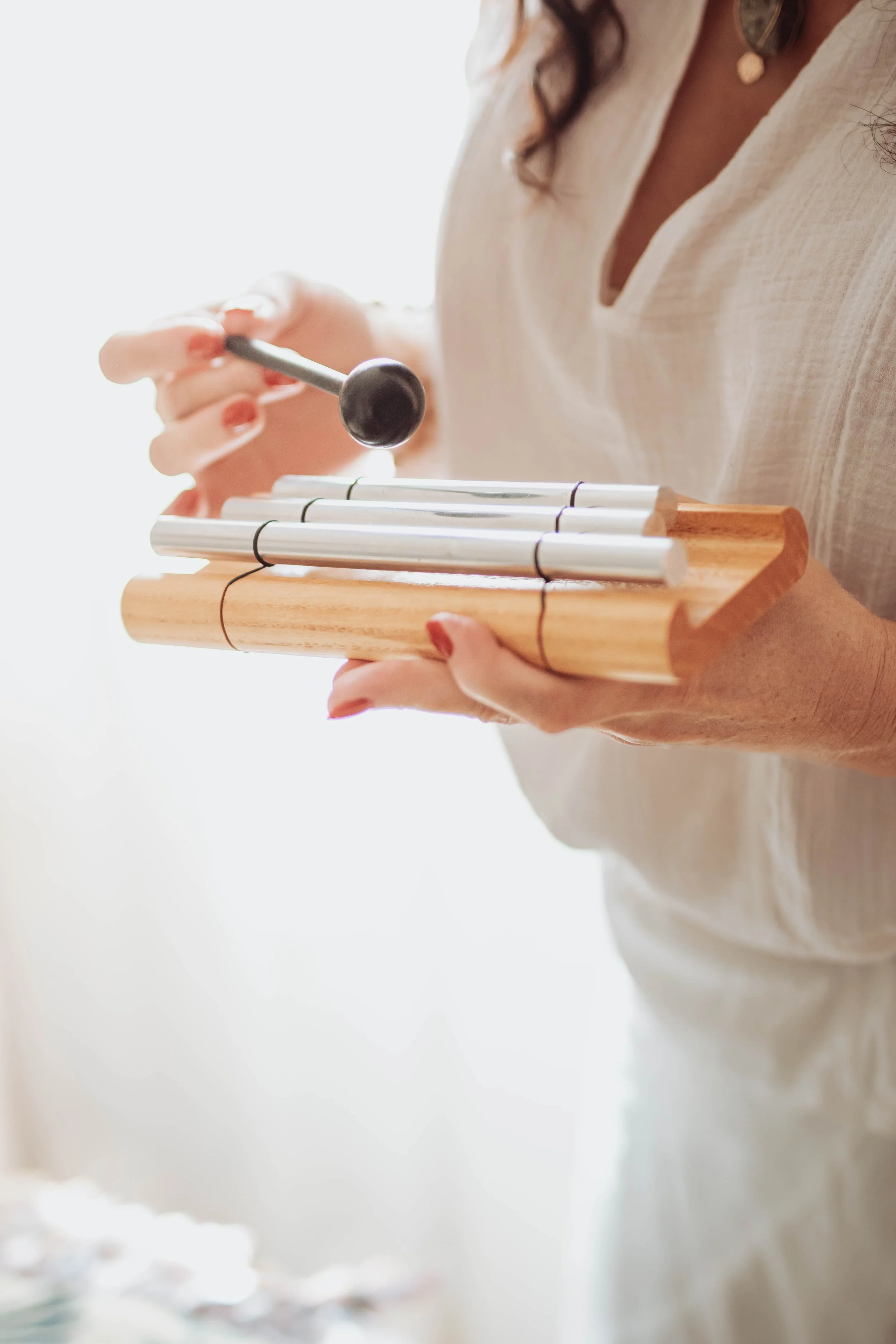 A person holding a small wooden xylophone with metal bars and a mallet, wearing a beige top and a necklace.