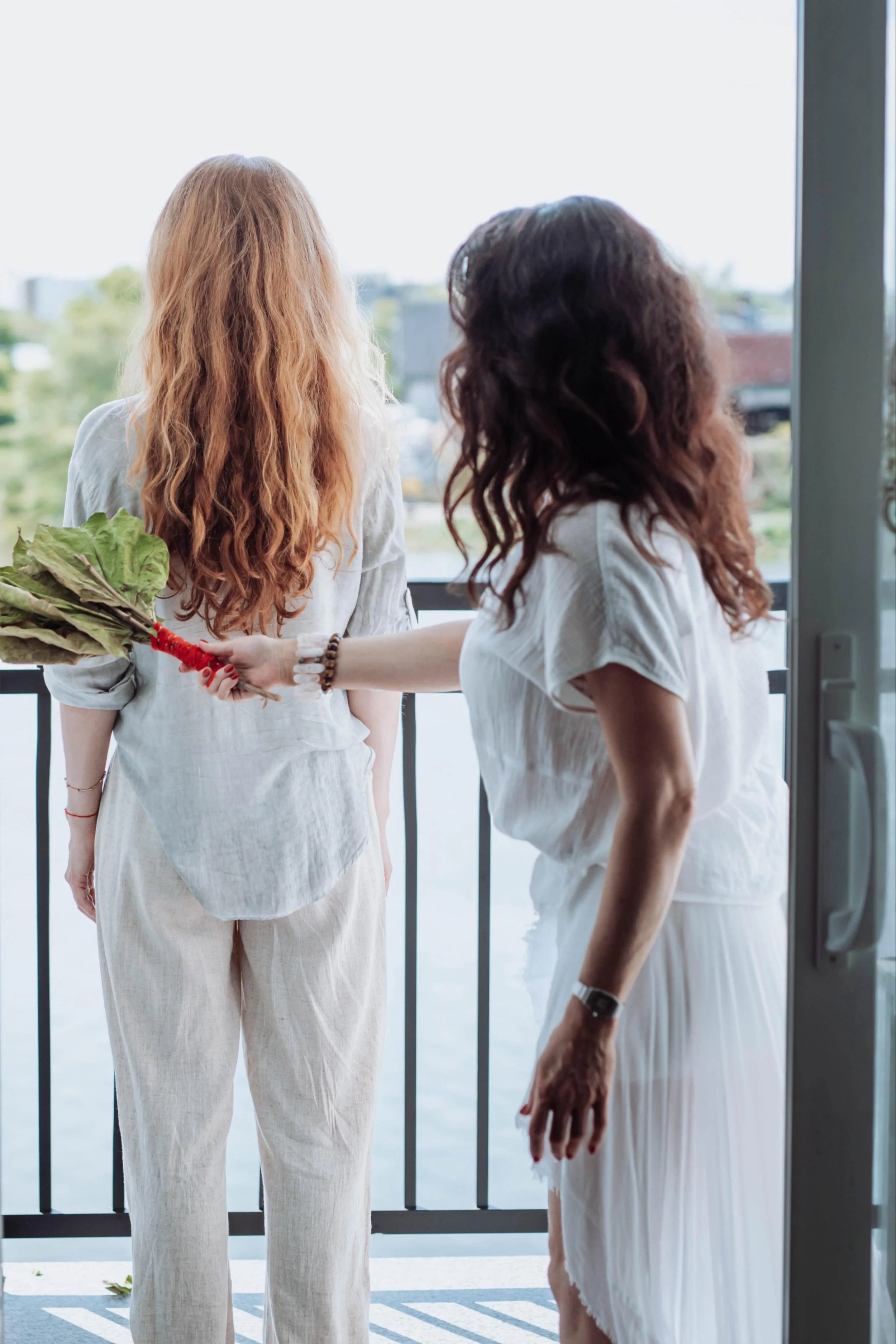 Two women standing on a balcony facing away, with one woman holding a bouquet of leafy greens. The scene is bright with a cityscape view in the background.