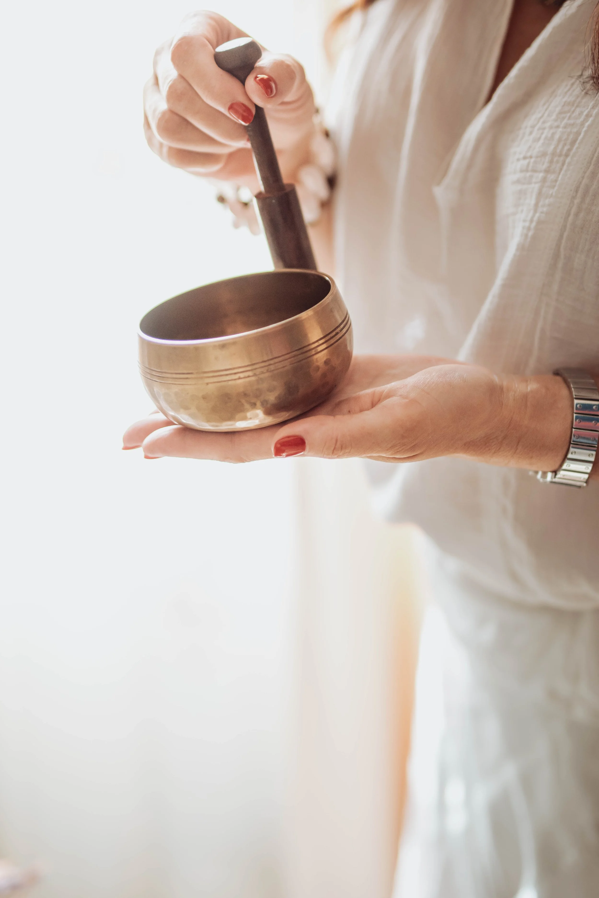 A person wearing a white shirt holding a singing bowl and a mallet, with part of their arm and wristwatch visible.
