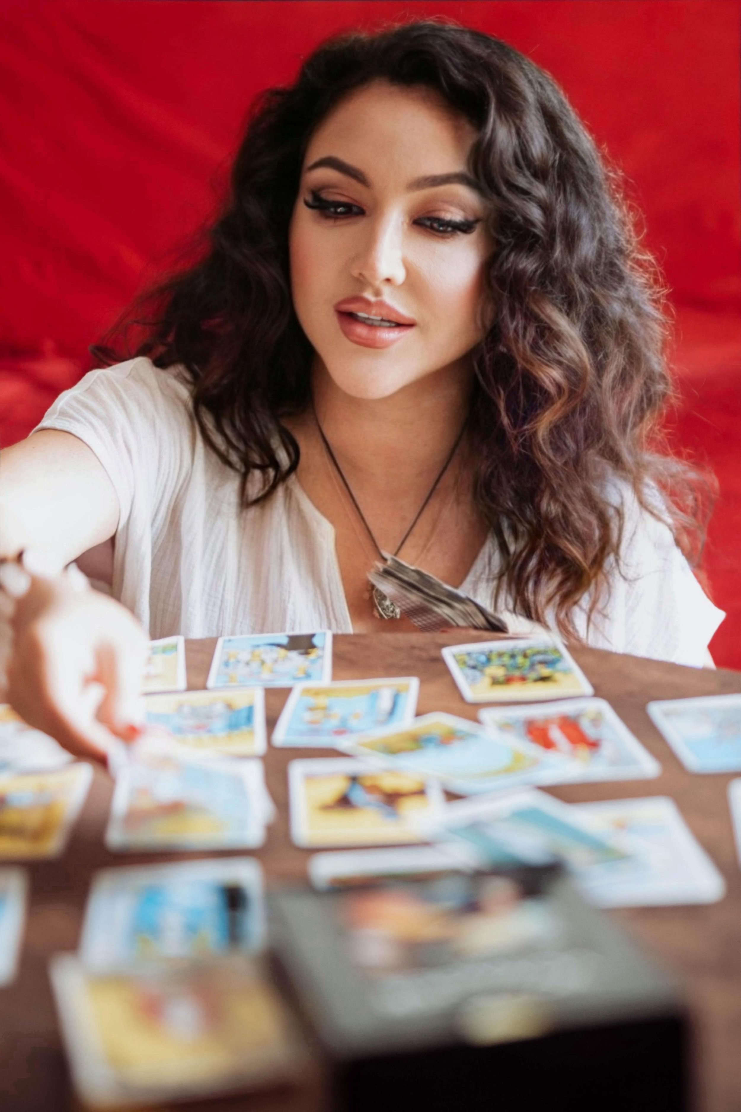 A woman with long, wavy brown hair, wearing a white top and a necklace, is looking at tarot or oracle cards spread out on a wooden table. The background features a red fabric or curtain.