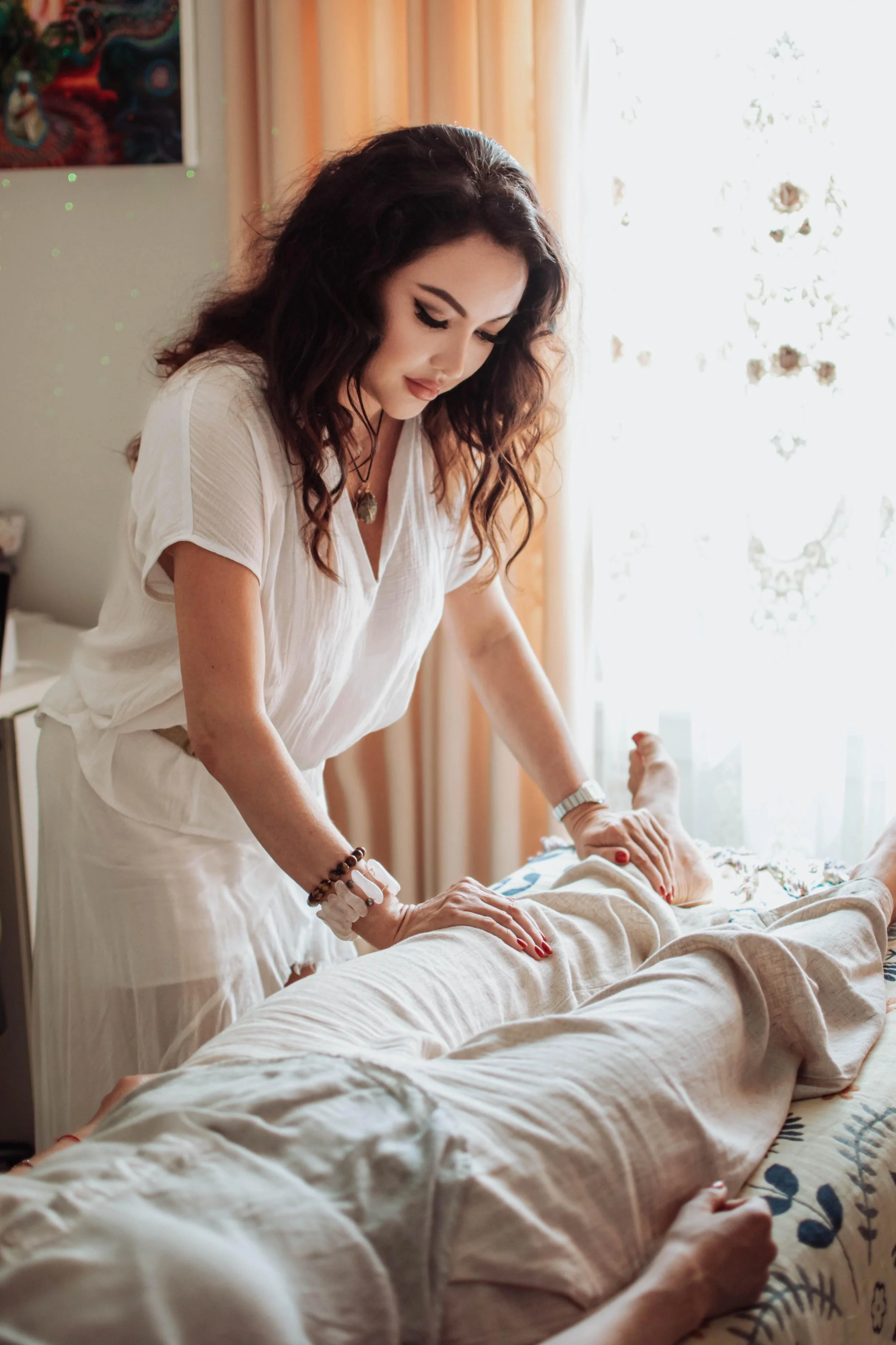 A woman with dark curly hair wearing a white top and jewelry is leaning over and caring for an elderly person lying on a bed. The elderly person is covered with a beige blanket, and their hand is visible, resting on the bed. The room has a bright window with floral curtains and a colorful painting on the wall.