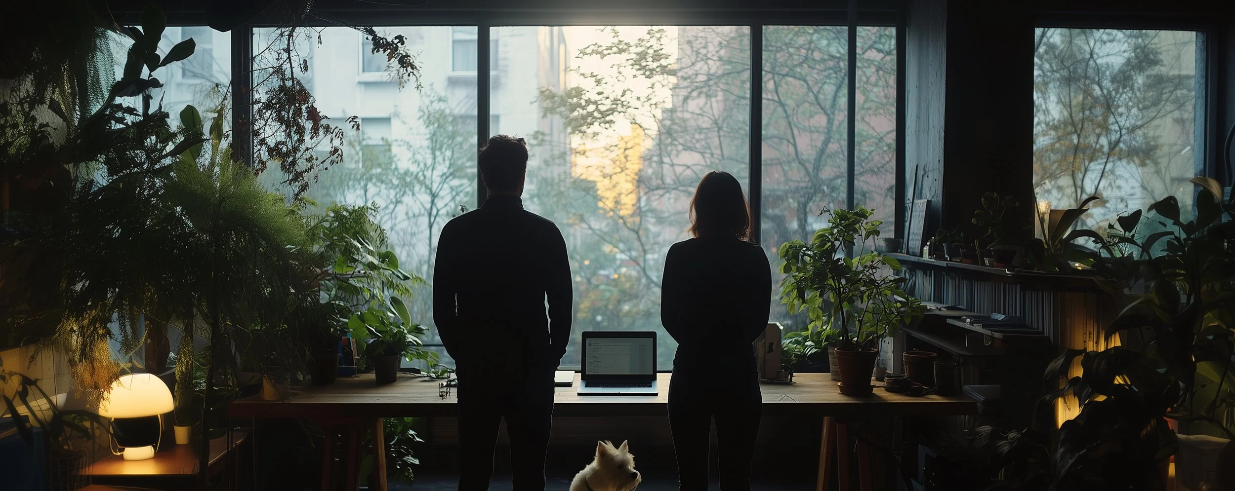 Dos personas mirando por una ventana grande en una sala llena de plantas, con una computadora portátil sobre una mesa. Hay un perro pequeño a sus pies.