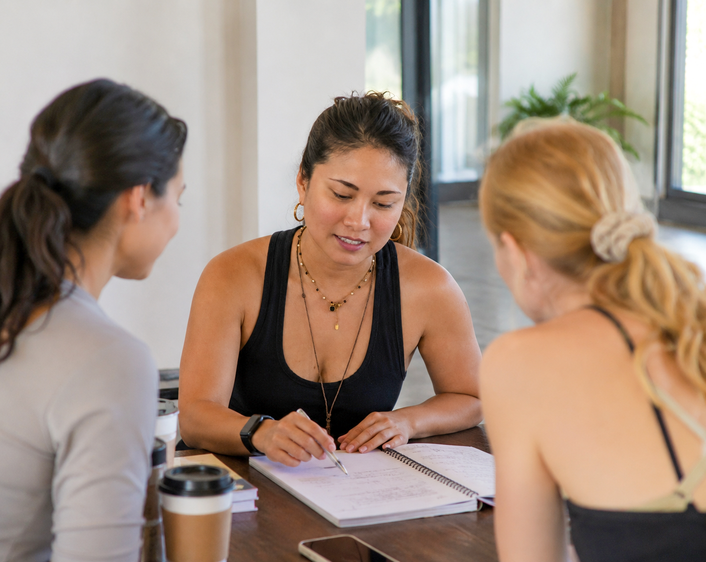 Three women sitting at a wooden table having a discussion. One woman is taking notes while the other two listen. There are coffee cups and a smartphone on the table.