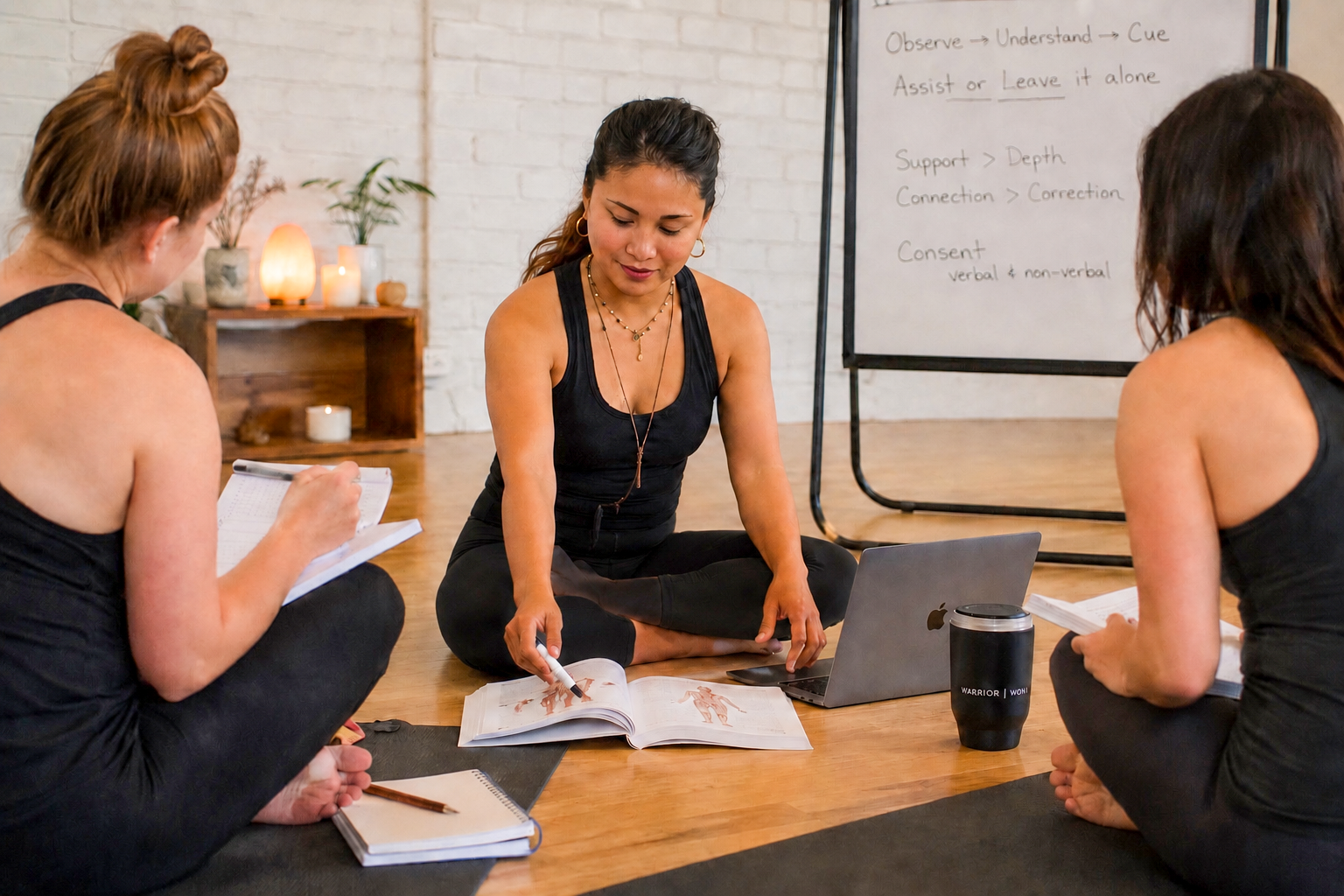 Three women sitting cross-legged on the floor during a yoga or meditation class, with notebooks, a laptop, and a whiteboard in the background.