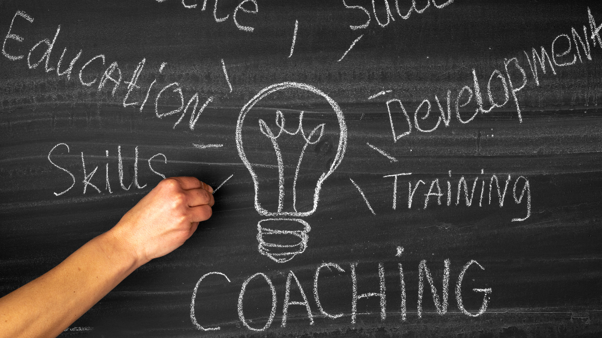 A blackboard with hand-drawn words related to skills development, training, and coaching, with a light bulb illustration at the center.