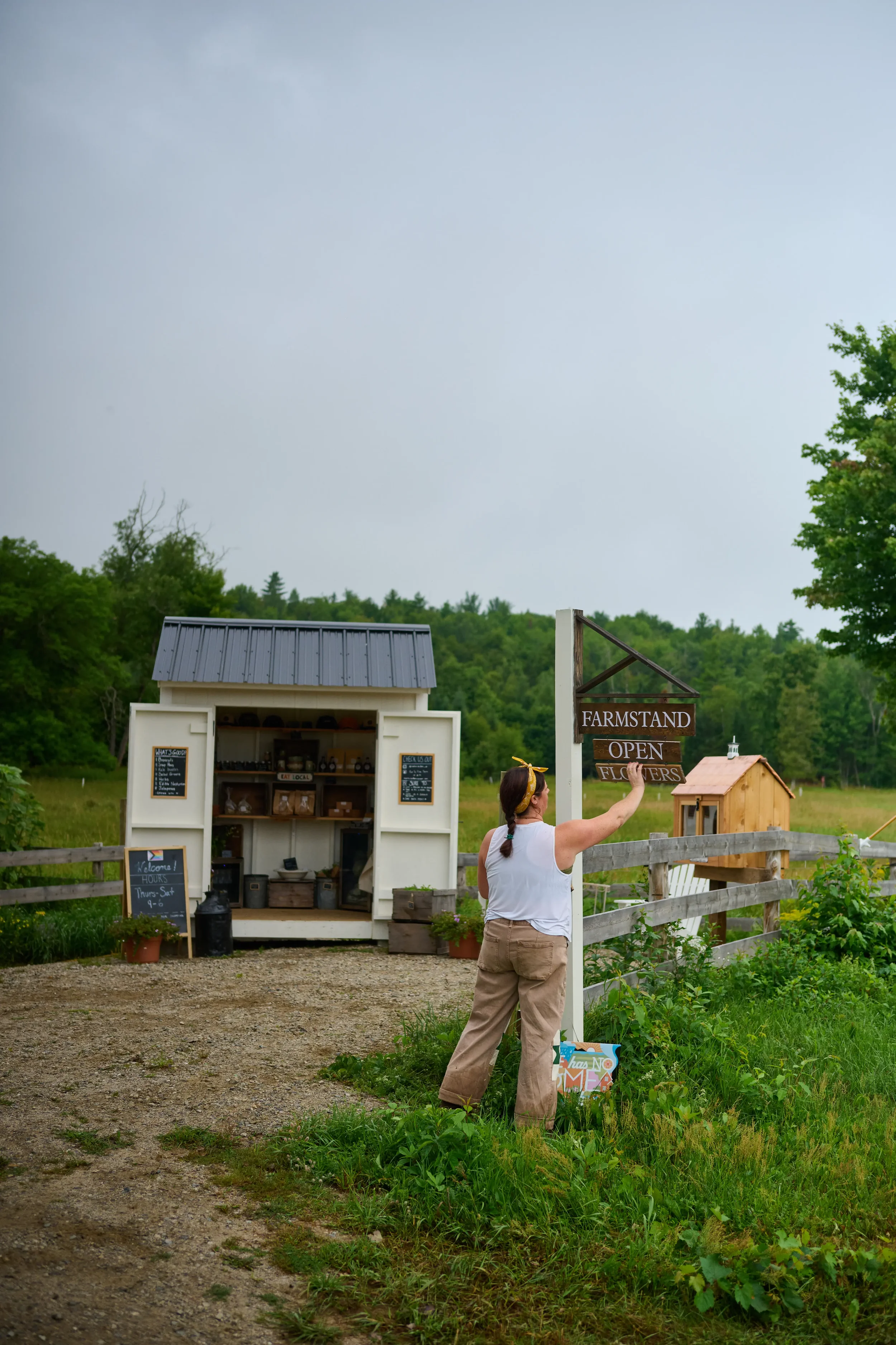 Woman standing outside near a farmstand sign, with a small white farmstand building in the background, surrounded by green trees and grass.