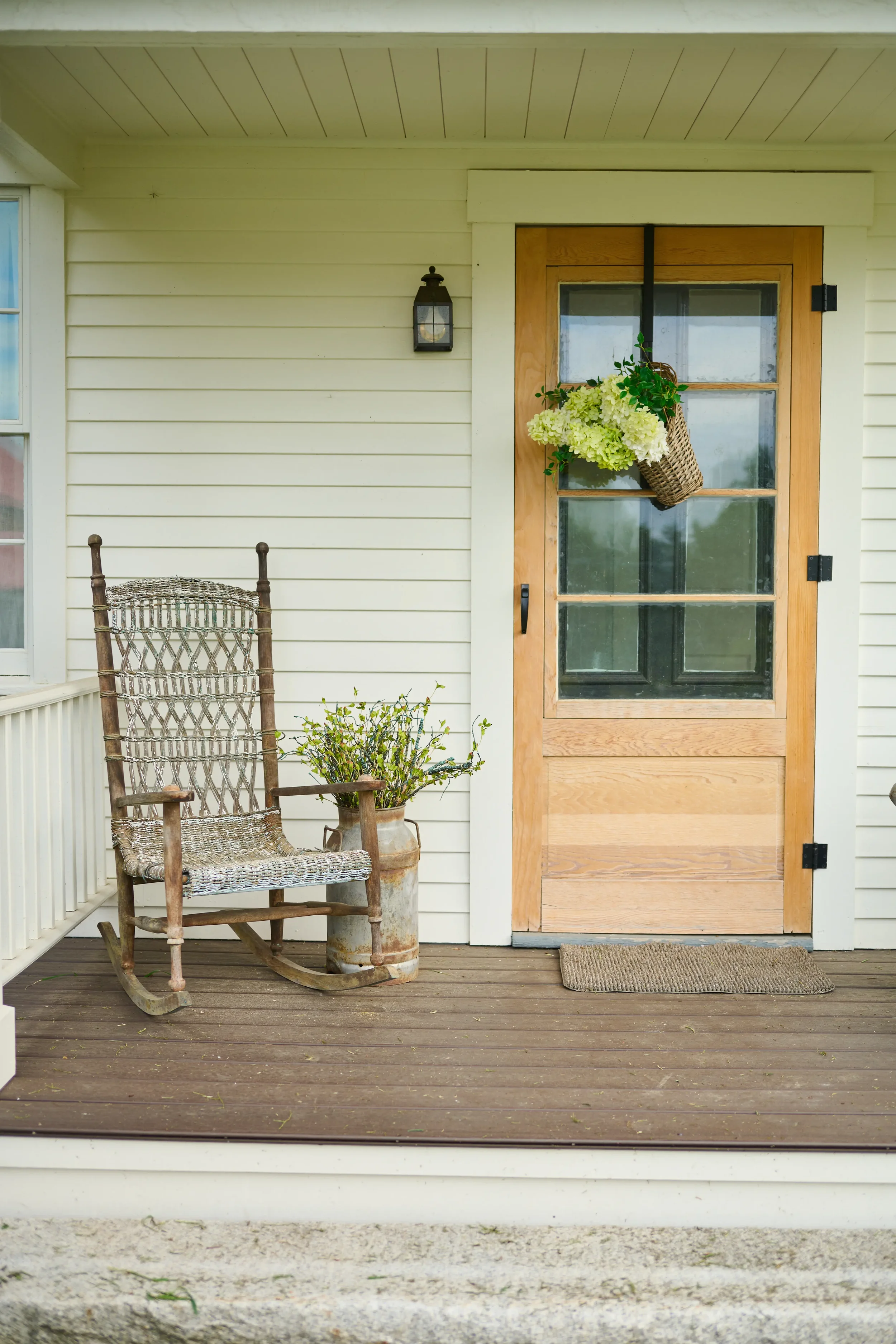A porch with a wooden rocking chair, potted plant, and a door with a hanging flower basket. Exterior siding is painted a light color, with a black lantern-style light fixture on the wall.