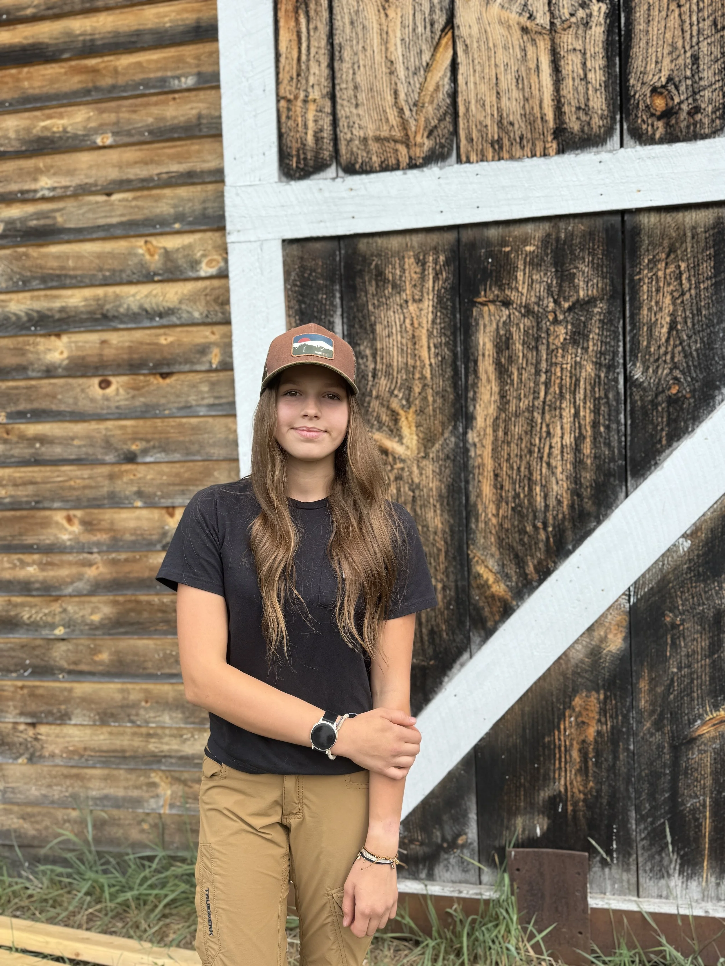 A young woman with long brown hair wearing a brown cap, a black T-shirt, and khaki pants standing in front of a rustic wooden barn wall.