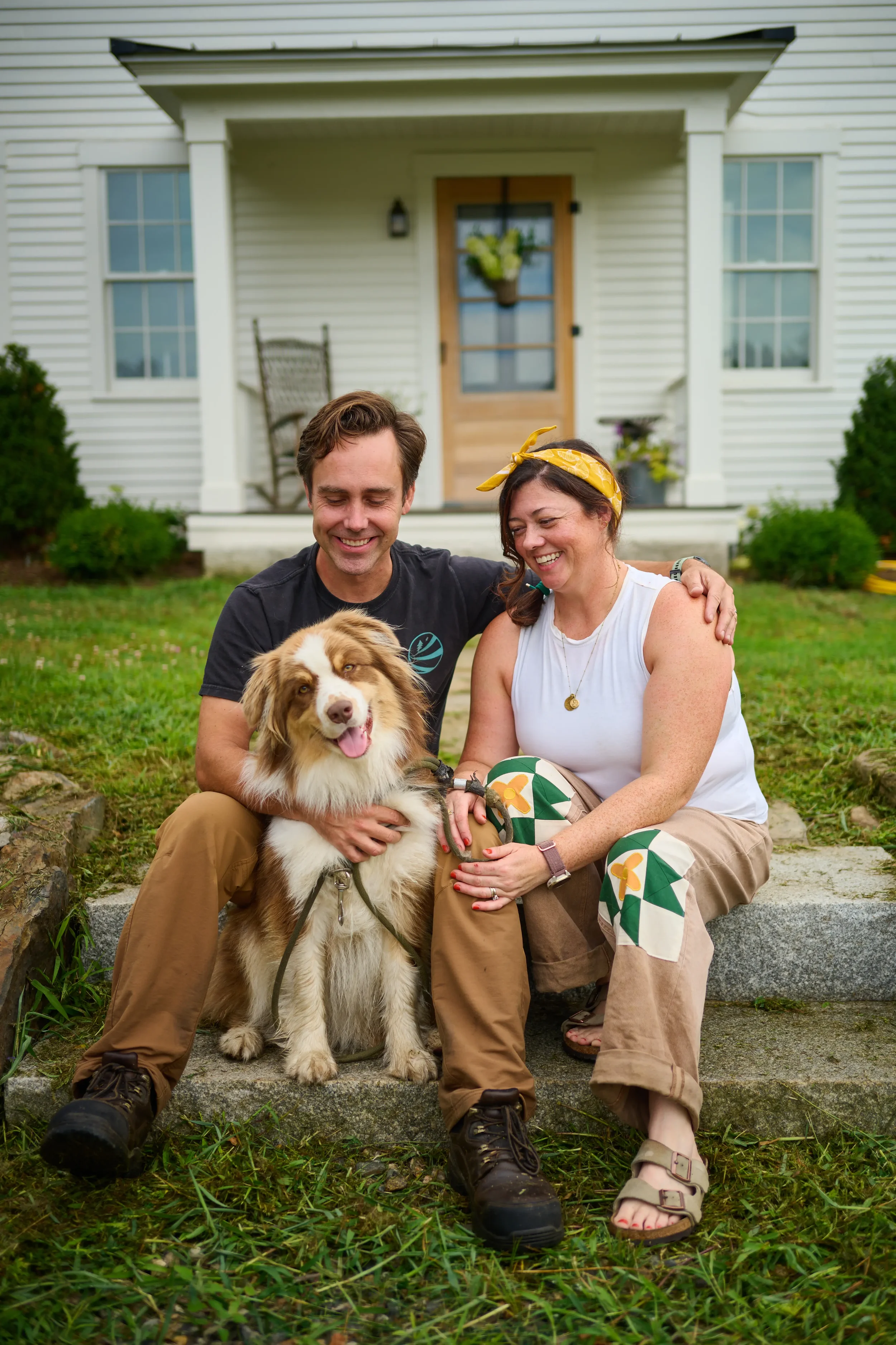 A happy man and woman sitting on steps outside a white house with a dog between them, smiling and cuddling, with greenery around.