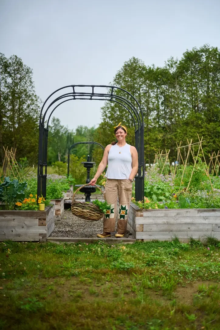 A woman in gardening clothes stands in a garden with raised beds, holding a basket of freshly picked vegetables, smiling.