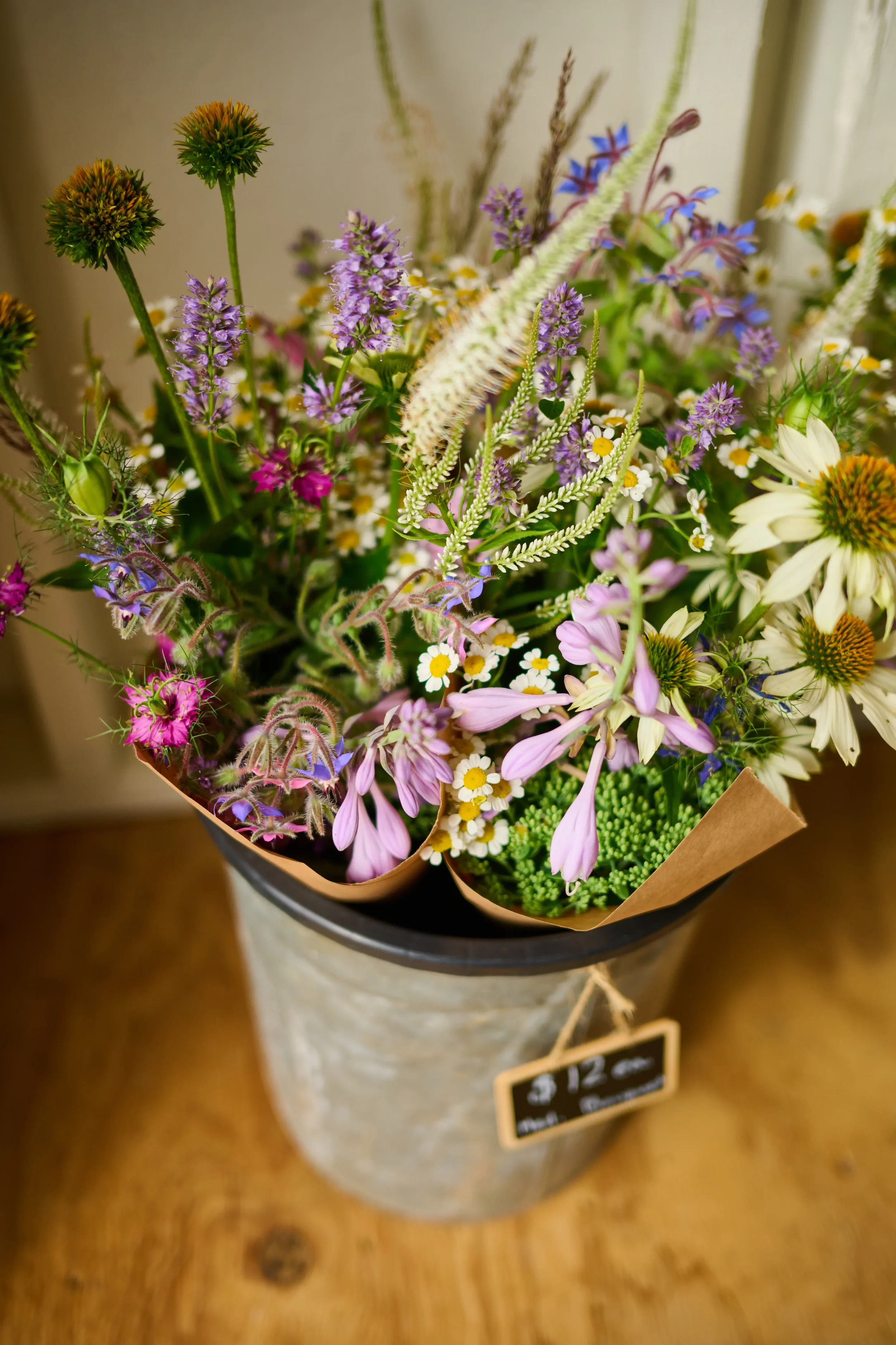 A bouquet of mixed flowers including purple, white, pink, and yellow blooms, arranged in a metal container with a brown paper wrapping.